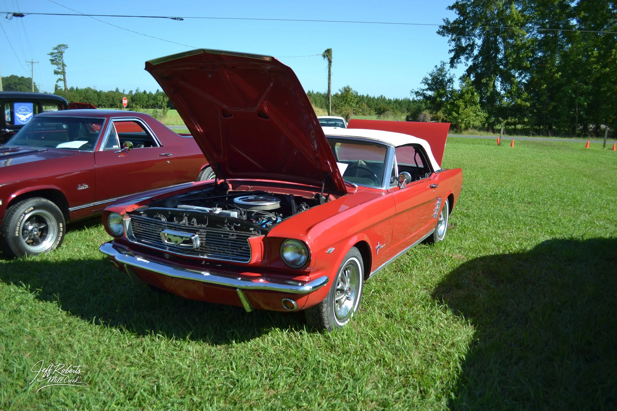 A red vintage Ford Mustang convertible with its hood open, parked on a grassy field at a car show. Next to it is a maroon classic car. Trees and orange traffic cones are visible in the background under a clear blue sky.