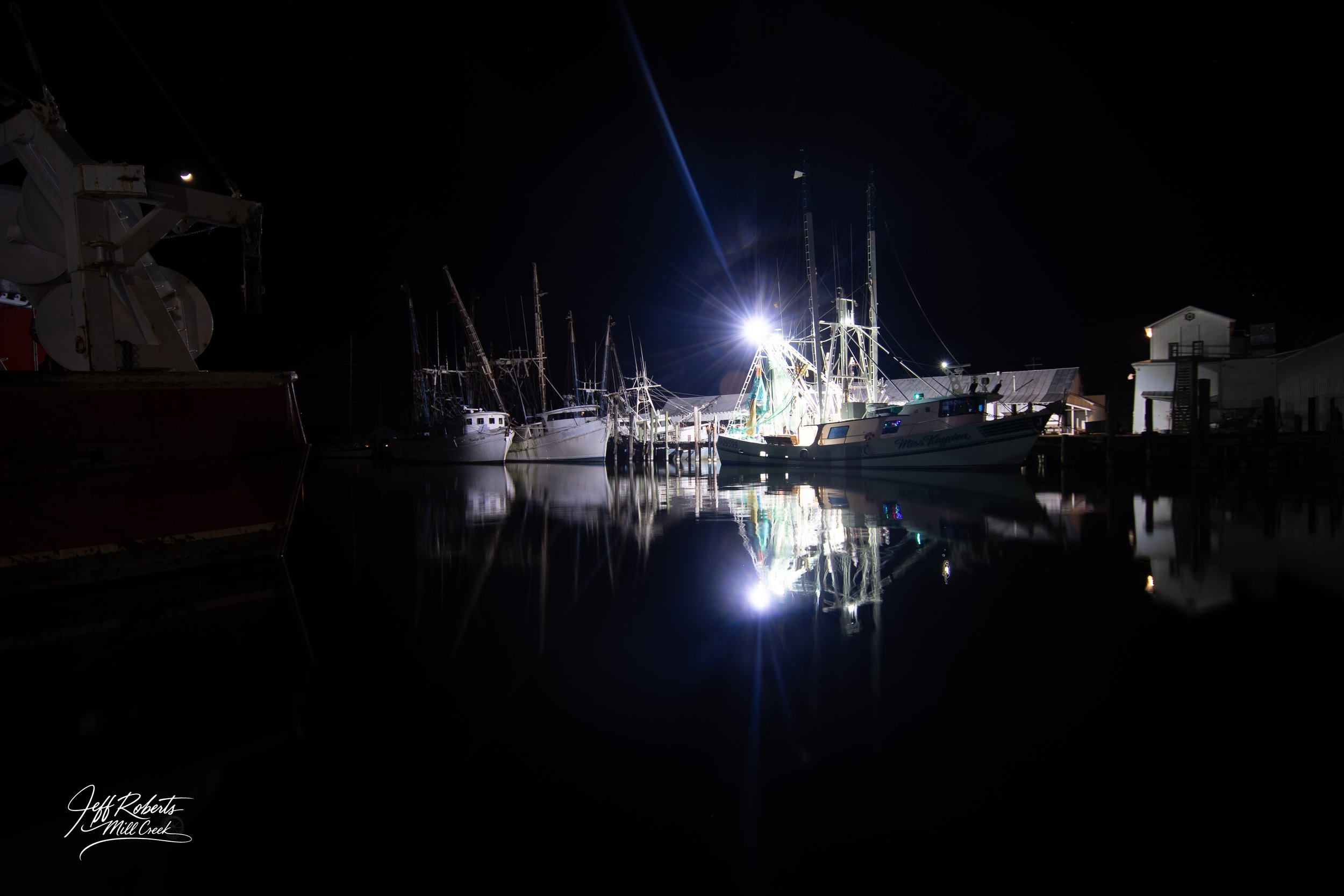 Nighttime scene of boats docked at a marina with bright light reflecting on the water, and buildings nearby.