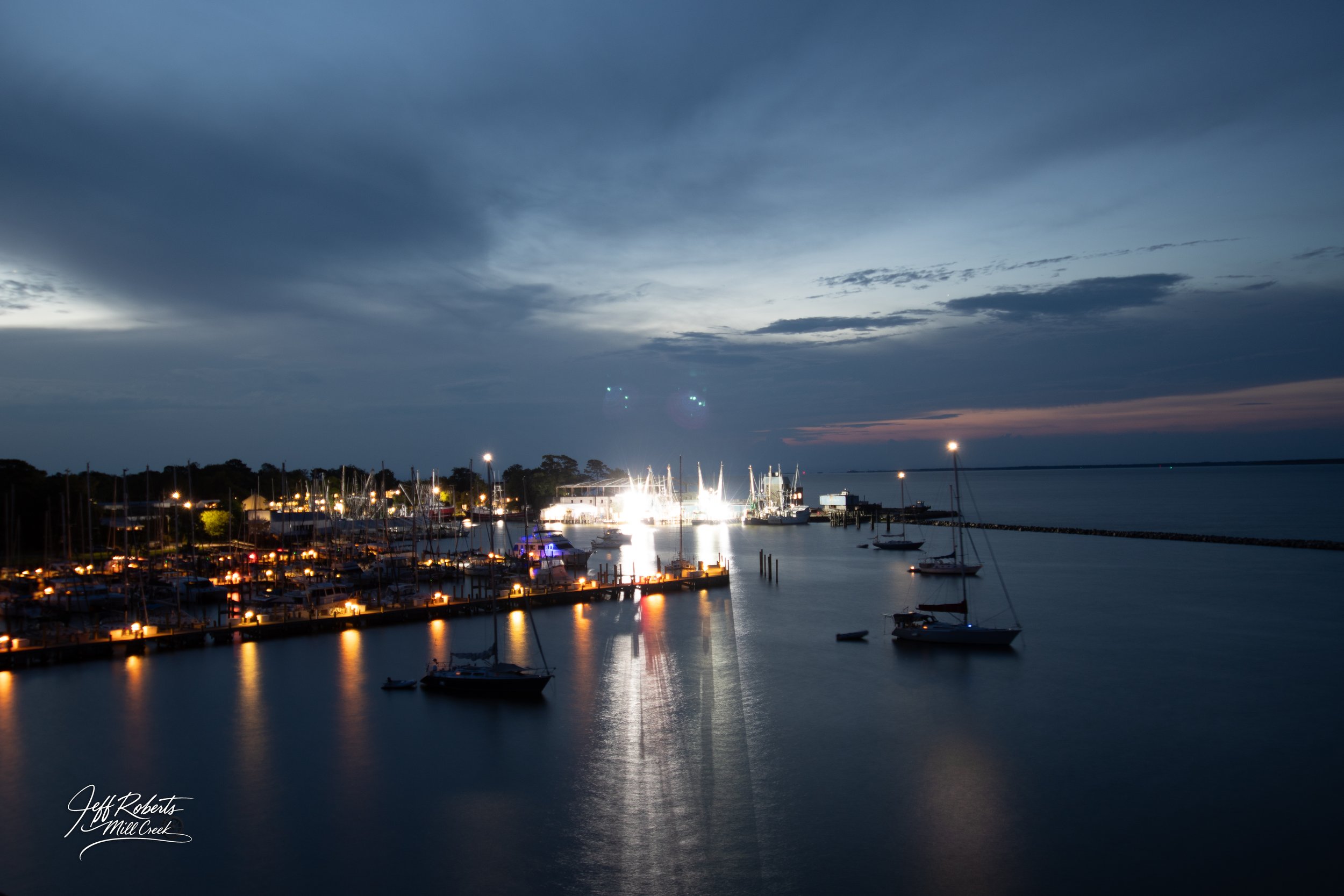 A peaceful marina at dusk with sailboats docked, some illuminated by lights, and a cloudy sky overhead.