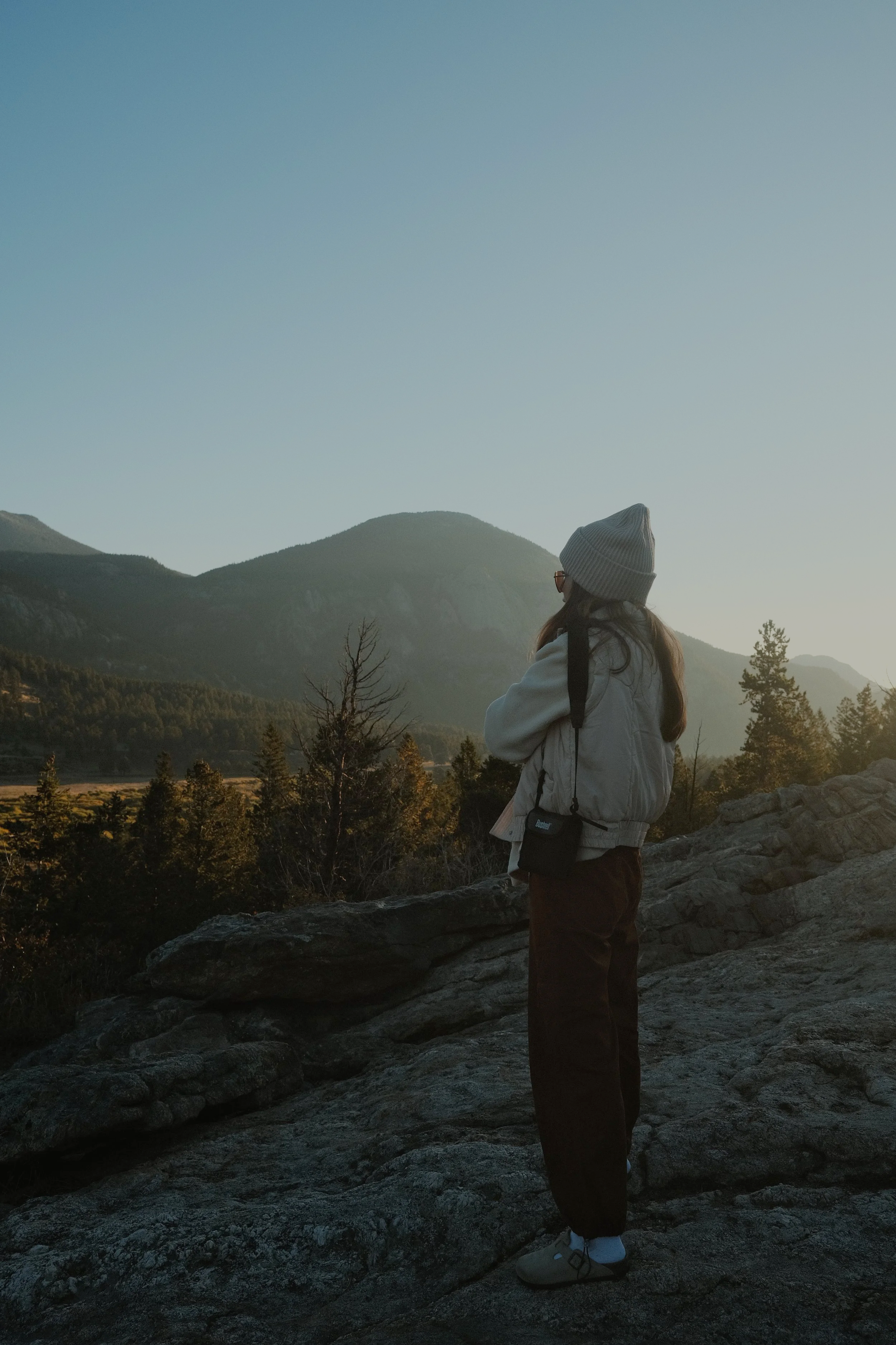 Person standing on a rocky overlook at sunrise, taking in a quiet mountain landscape with a sense of presence and intention.