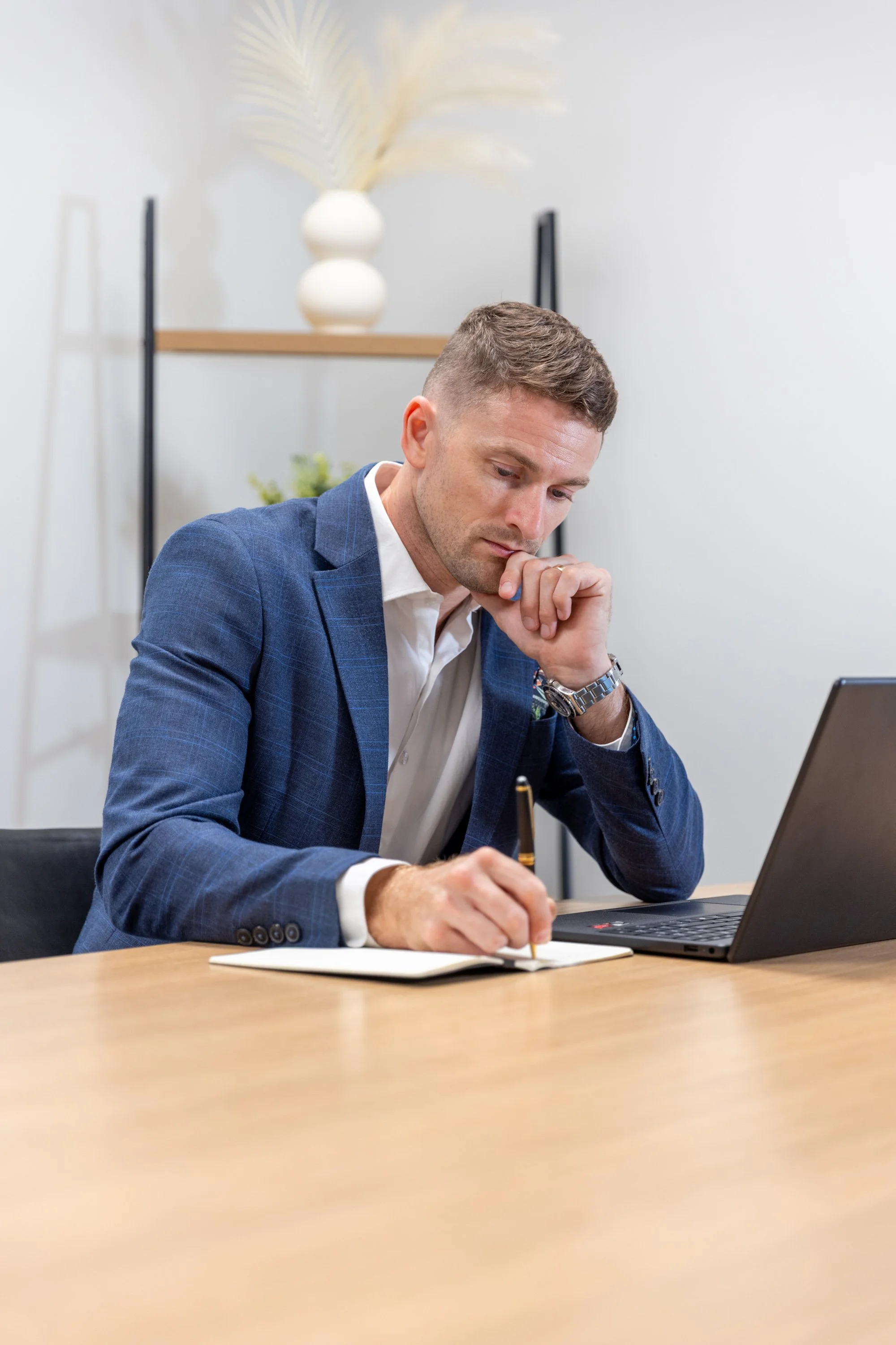 A man in a blue suit sitting at a desk, writing in a notebook with a pen, with a laptop open in front of him, in a modern office setting.