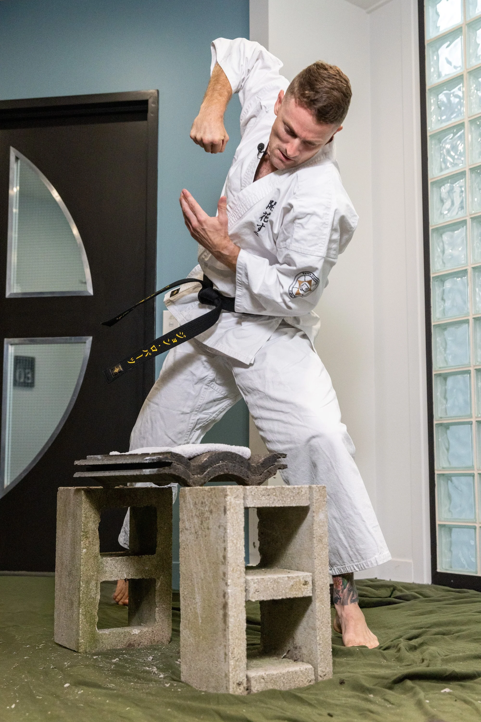 A man dressed in a white martial arts gi performing a martial arts move indoors, with cinder blocks stacked on the floor and a green mat underneath.
