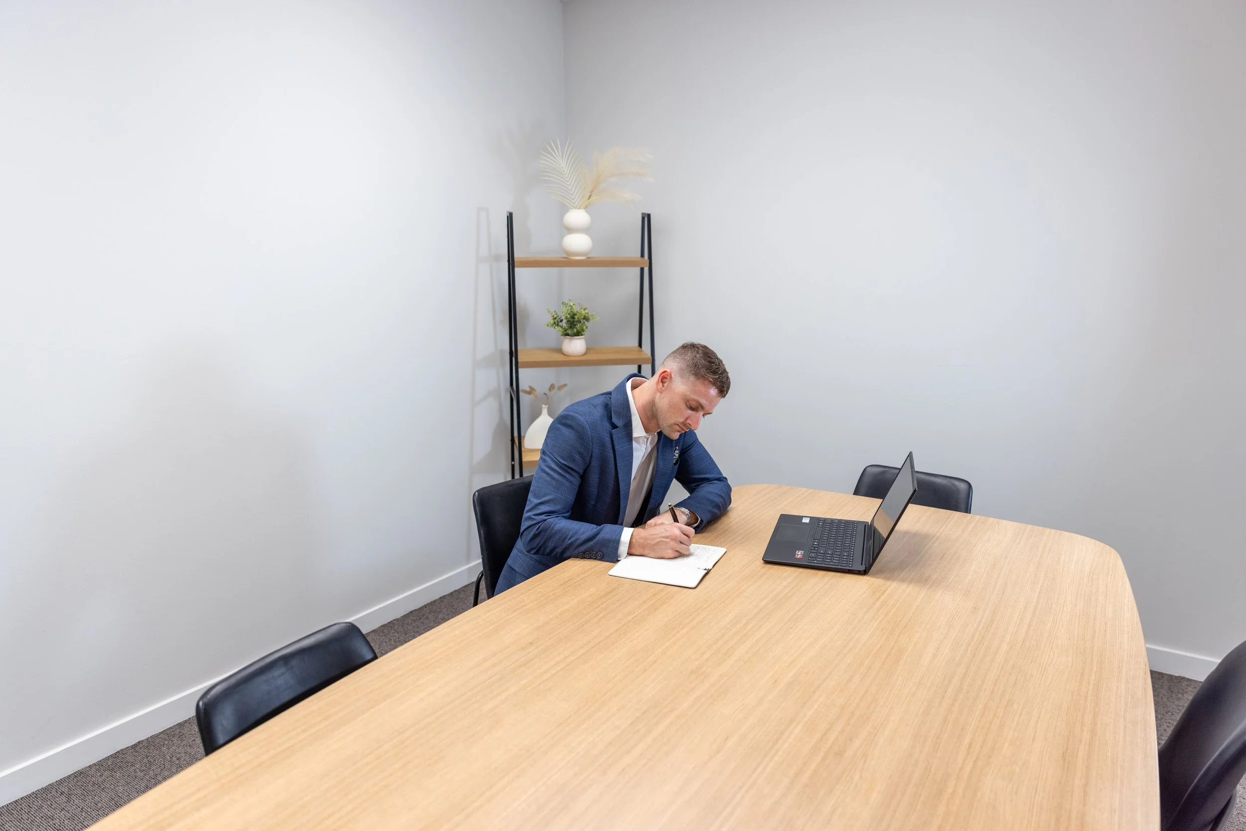 A man in a blue suit taking notes at a conference table in a minimalist office with white walls and a black metal and wood shelf with decorative plants.
