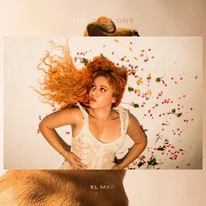 A young woman with curly, red hair poses with her hand on her hip in front of a white wall decorated with colorful confetti. She is wearing a white lace top and looking to the side with a playful expression.