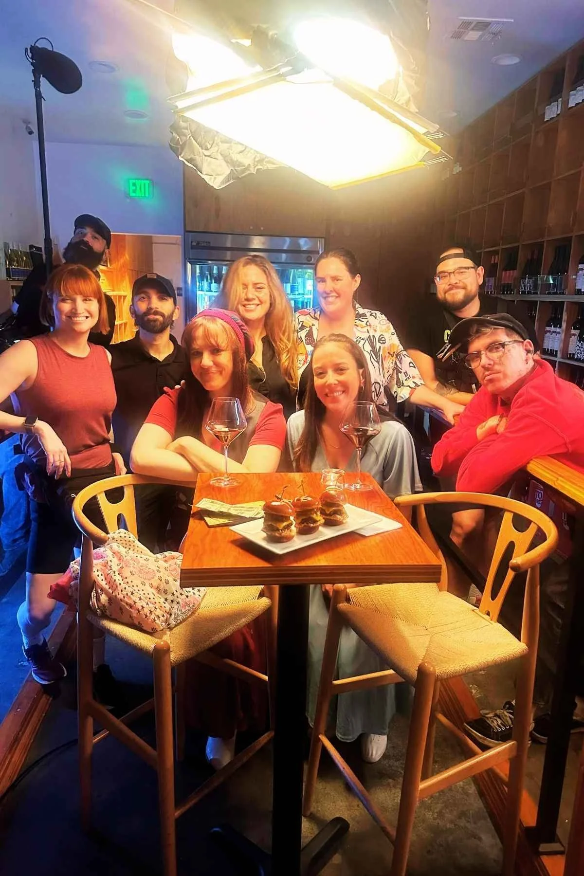 Group of nine people gathered around a table in a restaurant, with some holding glasses of wine, some smiling at the camera, and a plate of sliders on the table.