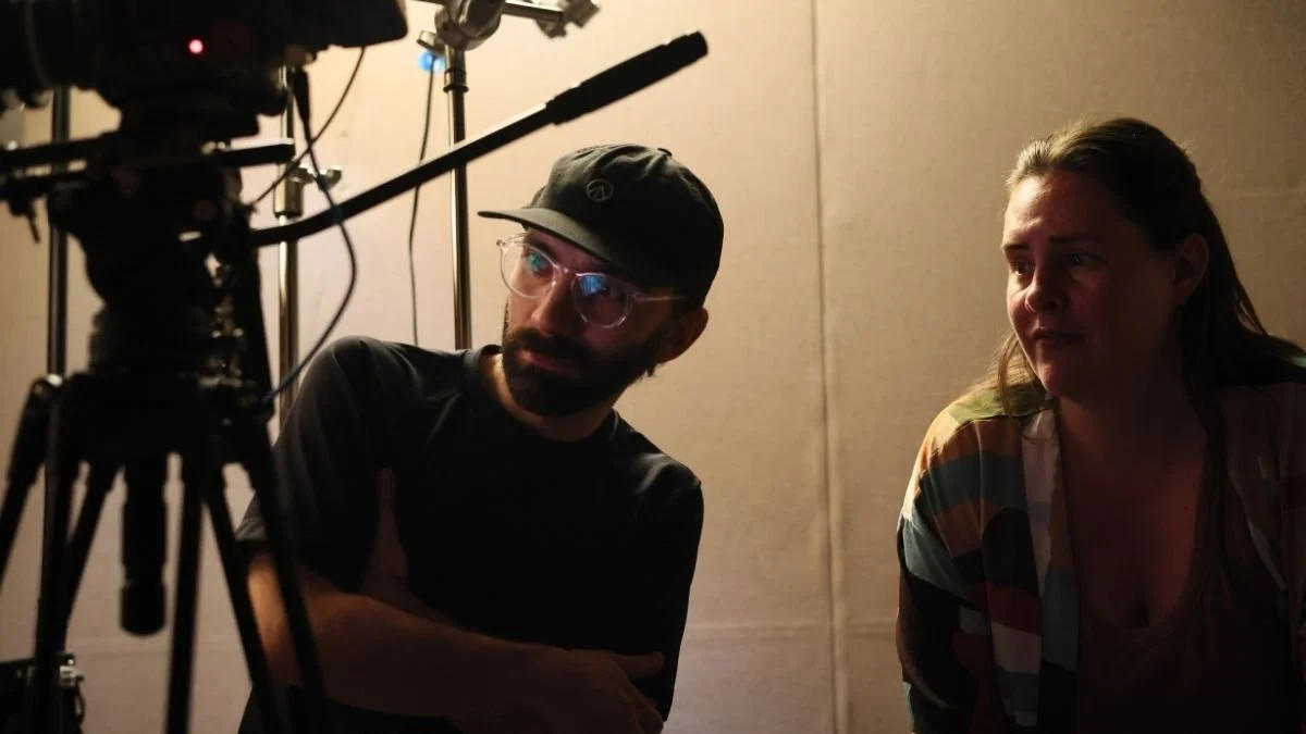 A man with glasses and a cap and a woman with long hair sit in a dimly lit room, looking at a camera or filming equipment.