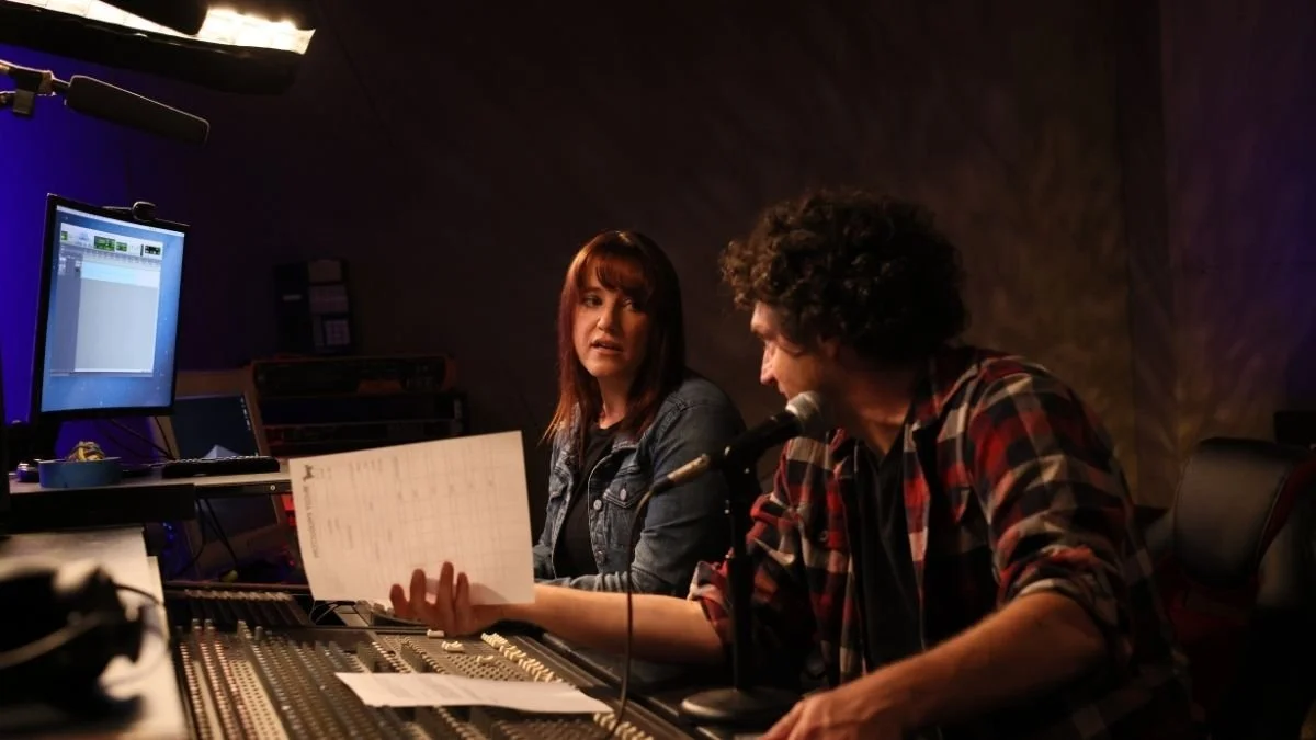 Two people in a recording studio, one man and one woman, sitting at a sound mixing console. The man with curly hair and a plaid shirt is holding a sheet of paper and speaking into a microphone, while the woman with straight hair and a denim jacket looks at him.