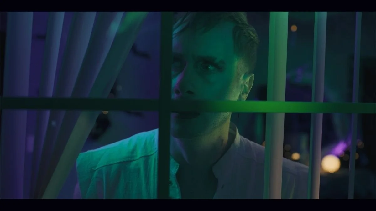 A young man with short hair looking through a window with bars, illuminated by colorful neon lights, at night.