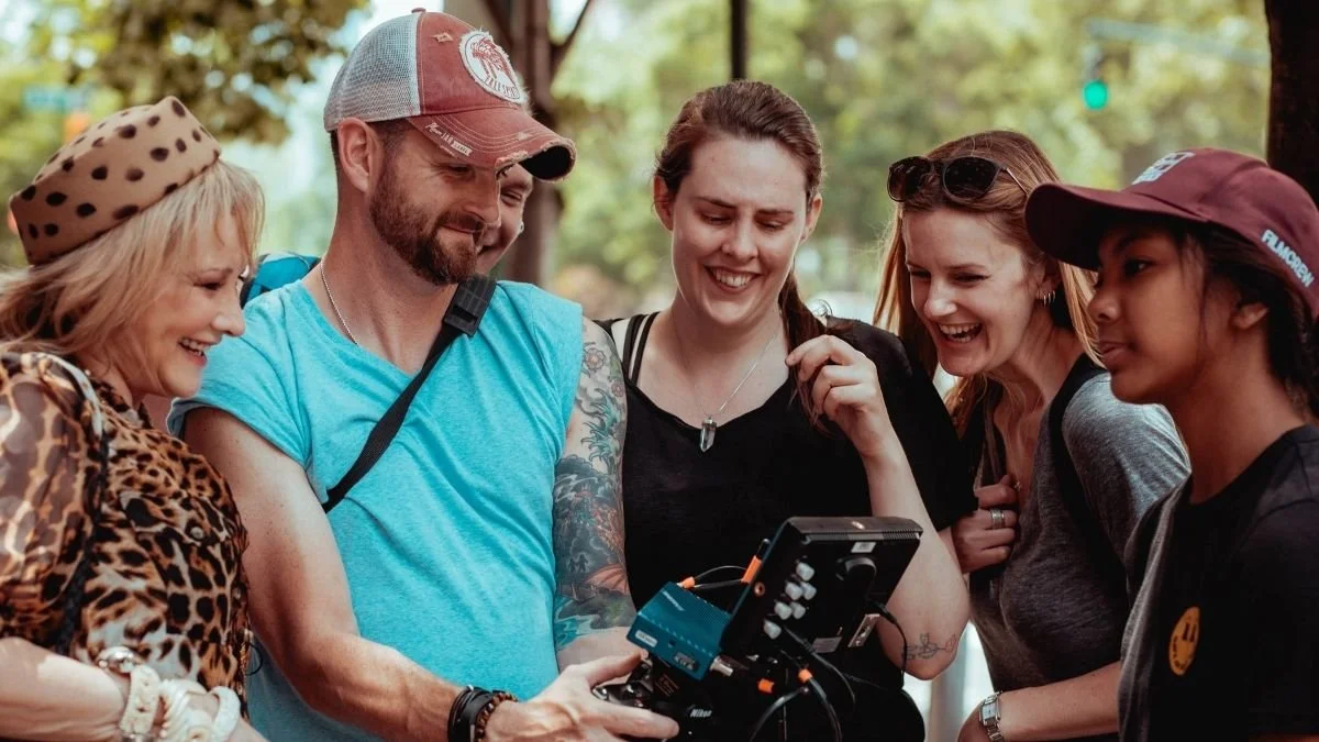 A group of six people, diverse in age and ethnicity, smiling and looking at a camera or device outdoors in a park.