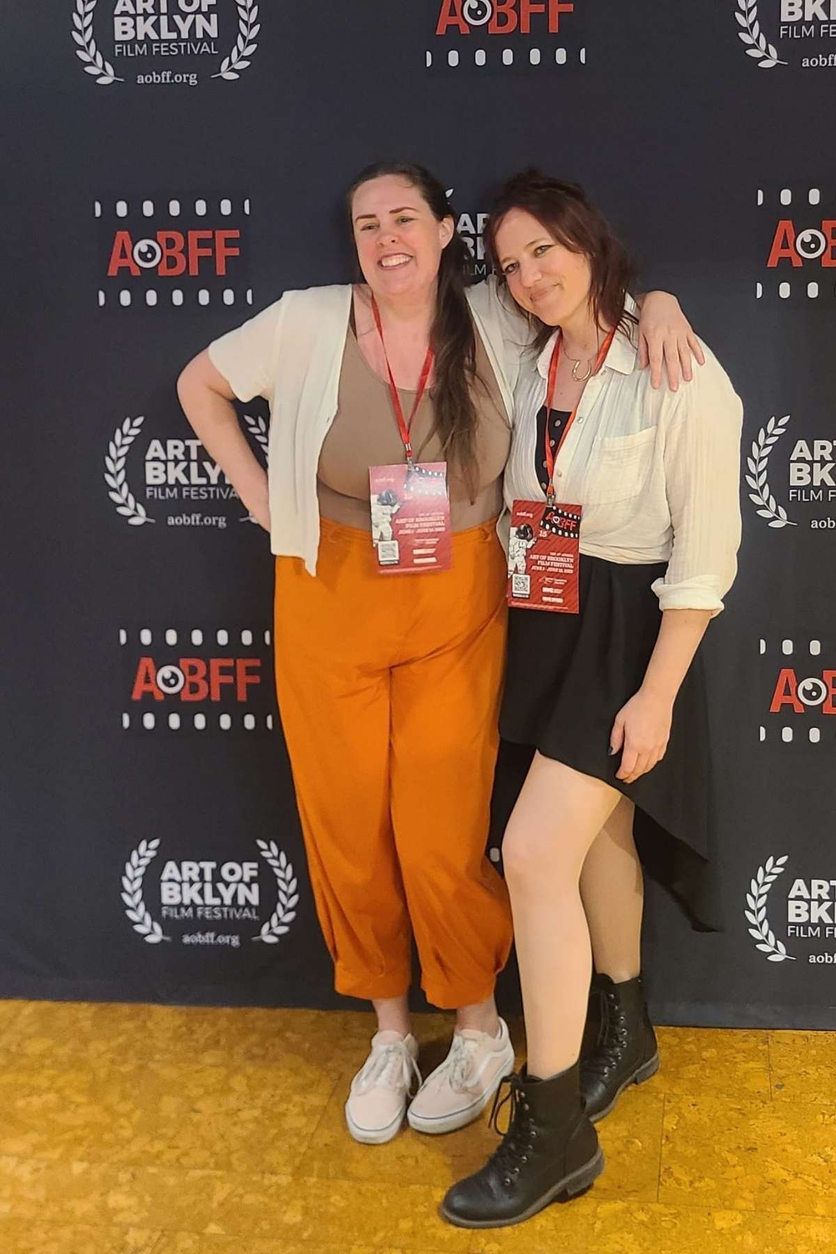 Two women standing together at the Art of Brooklyn Film Festival, smiling with arms around each other, in front of a black backdrop with festival logos and text.