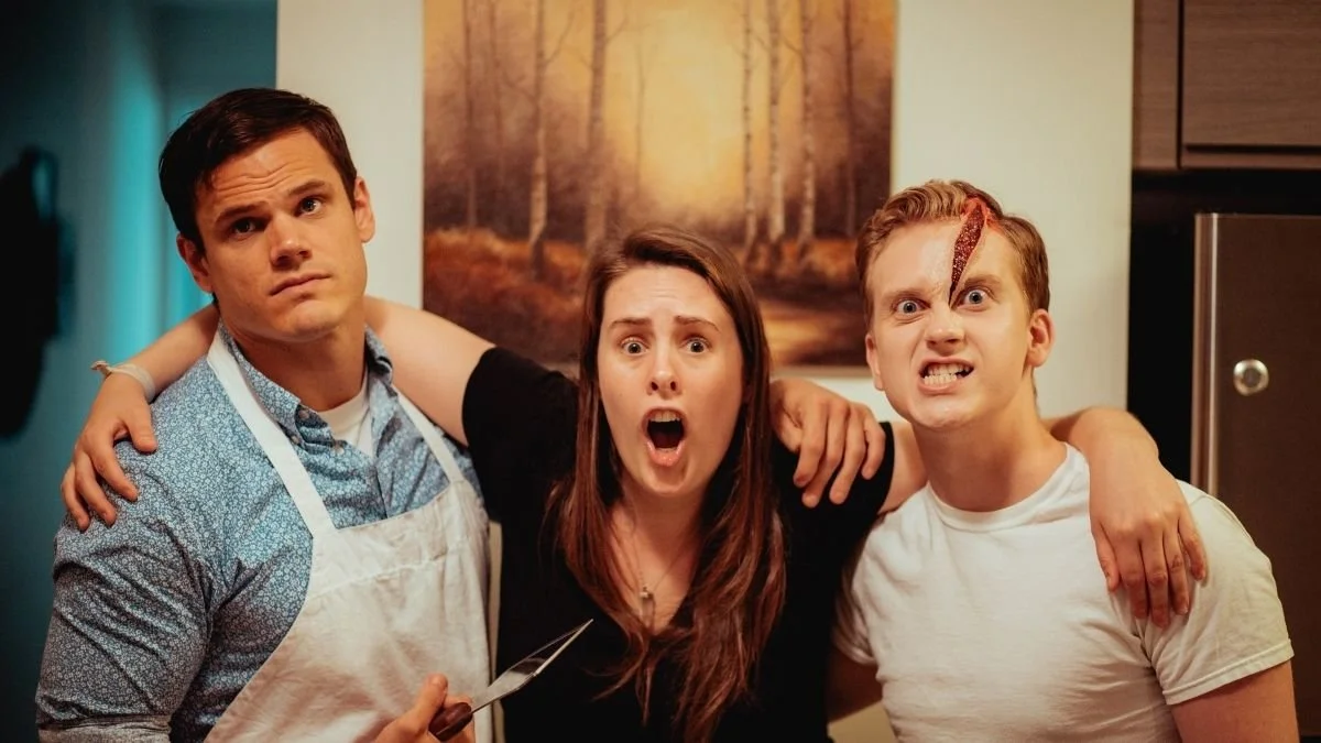 Three young adults with anxious and angry expressions, standing close together in a kitchen, with one holding a knife and a bandage on his forehead.