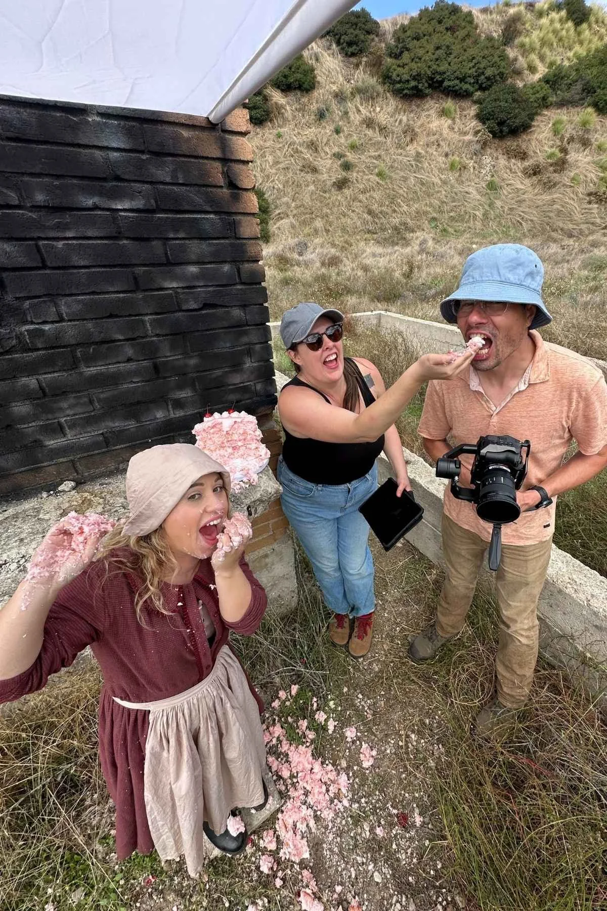 Three people are taking part in a pie-eating activity outdoors. One woman is feeding a man with a pie, while another woman, dressed in vintage clothing, is covered in pie and smiling. There are two pies on the ground and a pink cake with white frosting and cherries on top in the background.