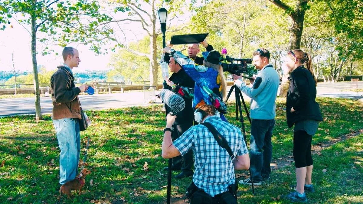 A young man holding a small dog and a blue object stands in front of a group of reporters and camera crew in a park with trees and a sidewalk.