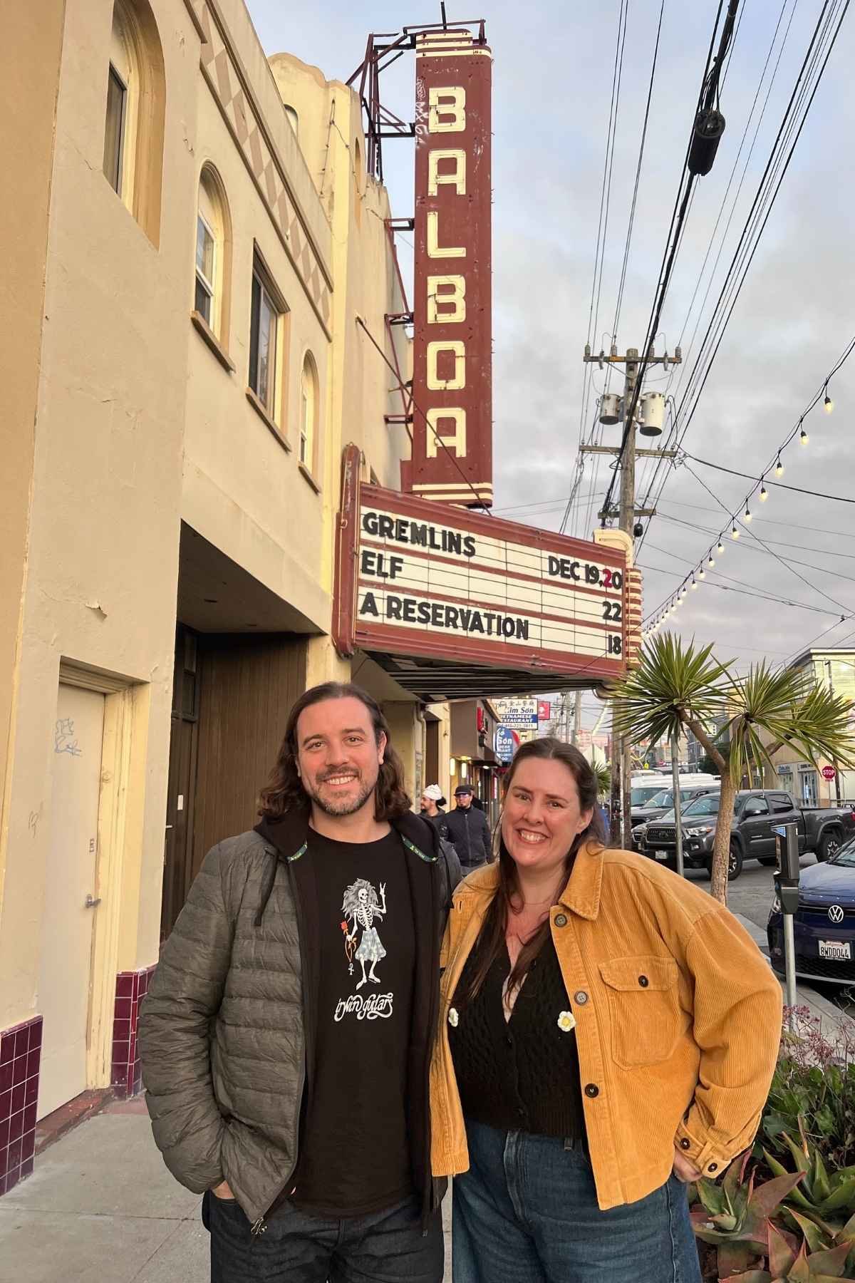 Two people standing outside a vintage theater with a marquee sign showing upcoming movies, smiling at the camera.