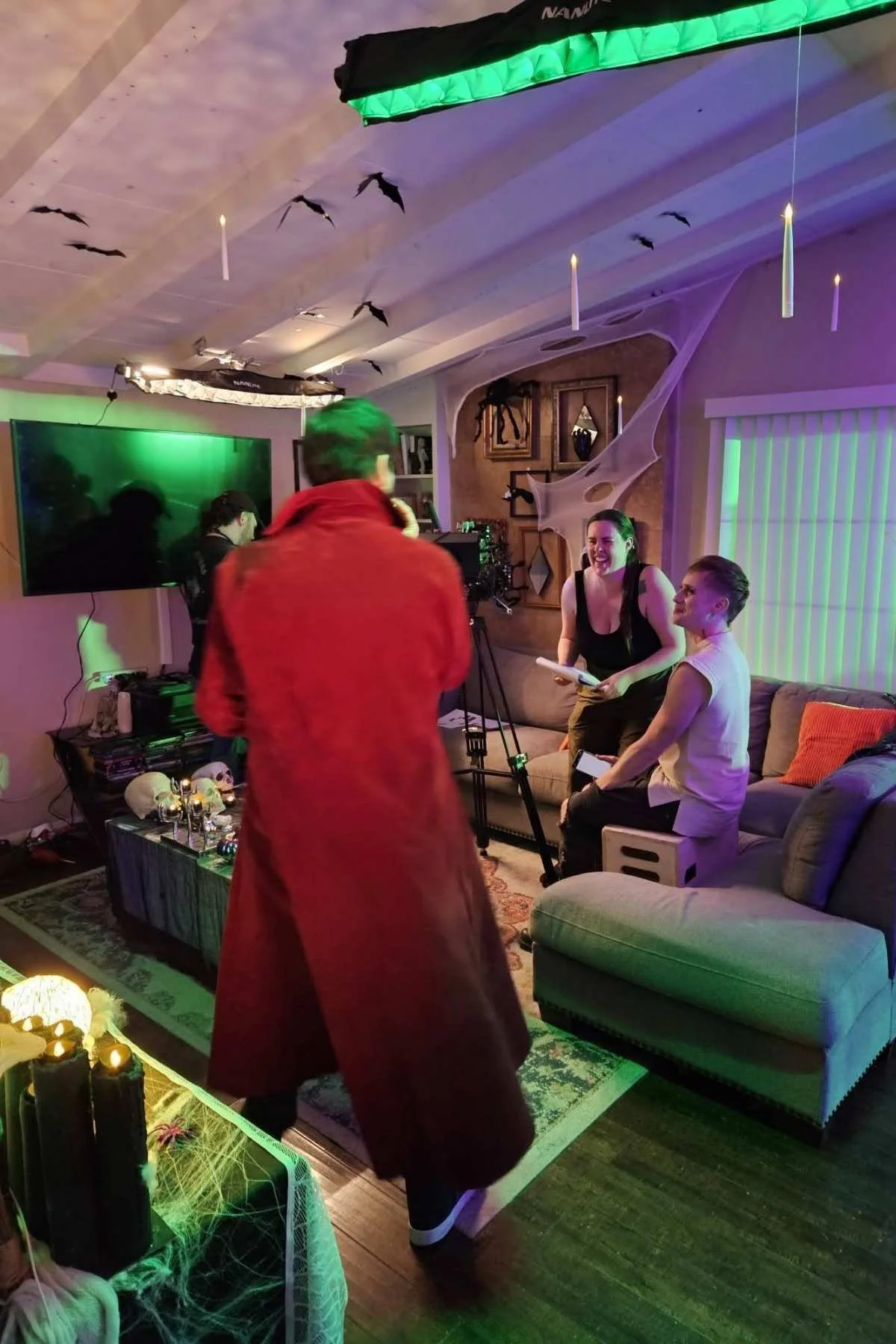 A Halloween-themed indoor scene with Halloween decorations, bat-shaped ceiling ornaments, spider web decorations, and Halloween candles with spider webs on a table. Four people are in the room: one man in a red coat with his back to the camera, two women sitting on a couch talking, and one person working with a camera.
