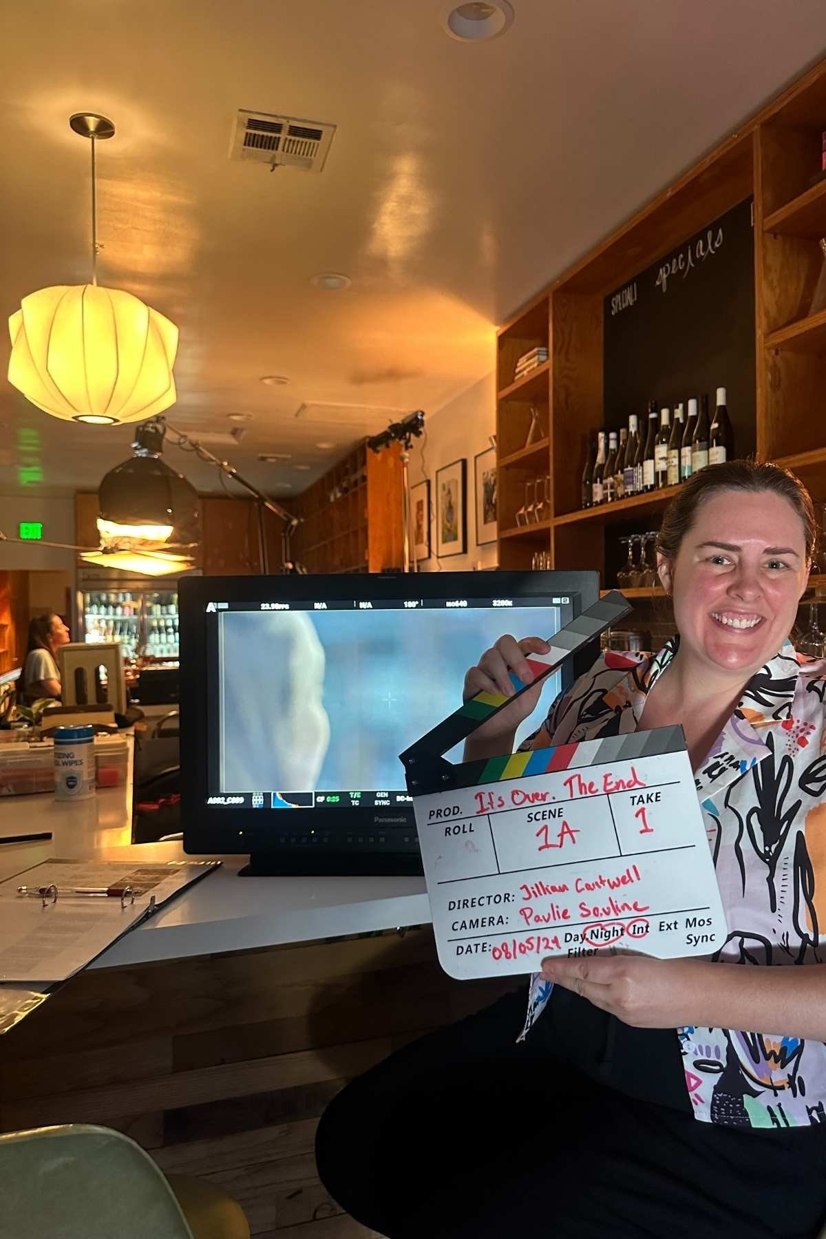 A woman smiling while holding a clapperboard in a restaurant or bar. Behind her, a monitor displays a close-up image. The background shows shelves with bottles of wine and barware, warm lighting, and framed pictures on the wall.