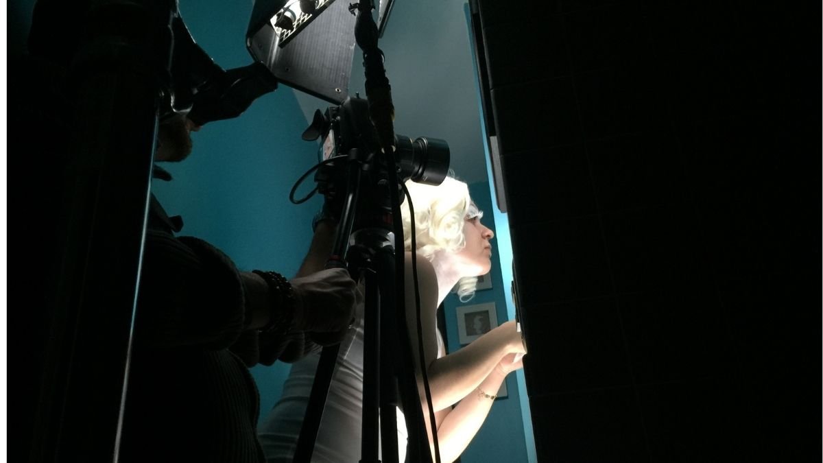 A person with blonde hair and glasses sitting at a desk, looking intently at a monitor, with a camera and technician filming or photographing them in a dimly lit room with a blue wall.