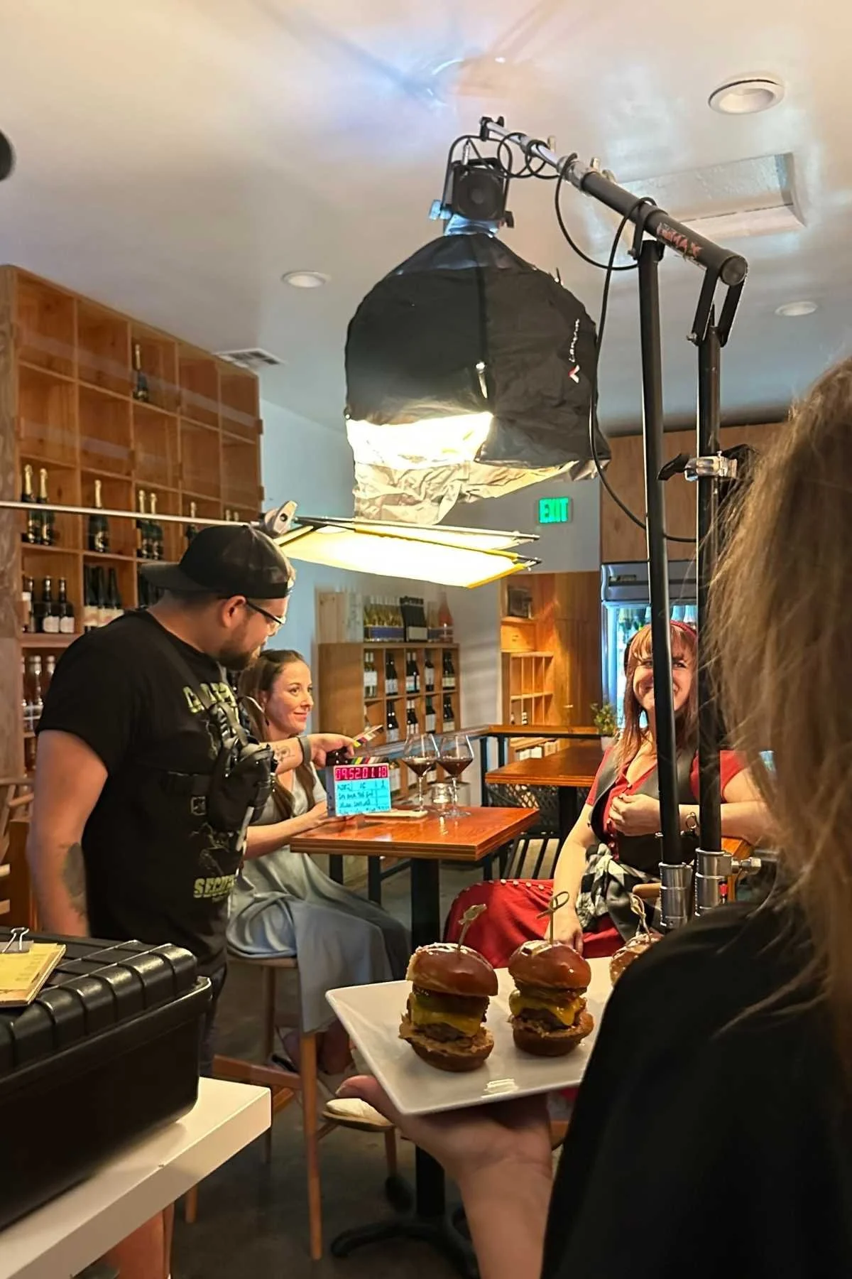 A behind-the-scenes view of a filming set in a restaurant environment with three women at a table and a man with a camera. The woman in the foreground is holding a tray with three mini burgers, and there is lighting equipment overhead.