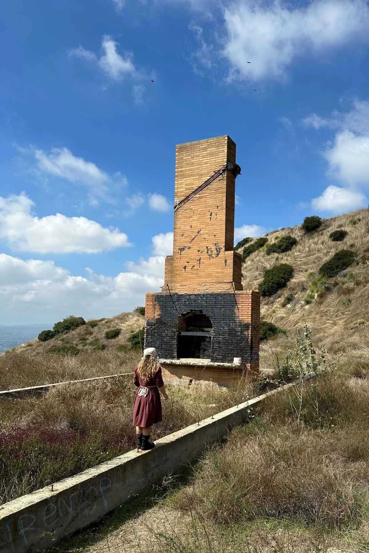 A woman in a period costume observing a large, dilapidated brick outdoor oven or furnace on a grassy hillside under a partly cloudy blue sky.
