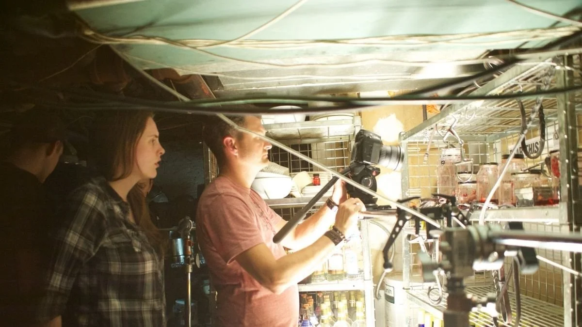 Two women and a man behind-the-scenes on a film set, with camera equipment and lighting, in a room with shelves of bottles.