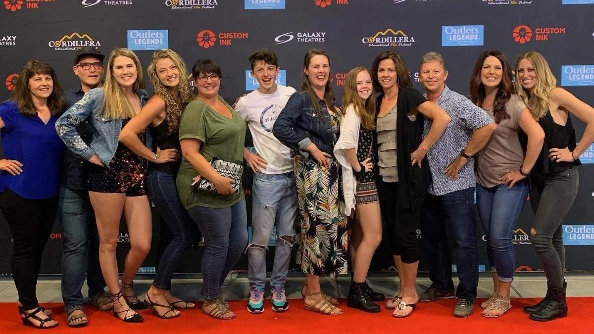 Group of fifteen people standing on a red carpet, smiling, at an event with a dark backdrop featuring various logos including 'Galaxy Theatres', 'Outlets Legends', 'Custom Ink', and 'Cordillera'.
