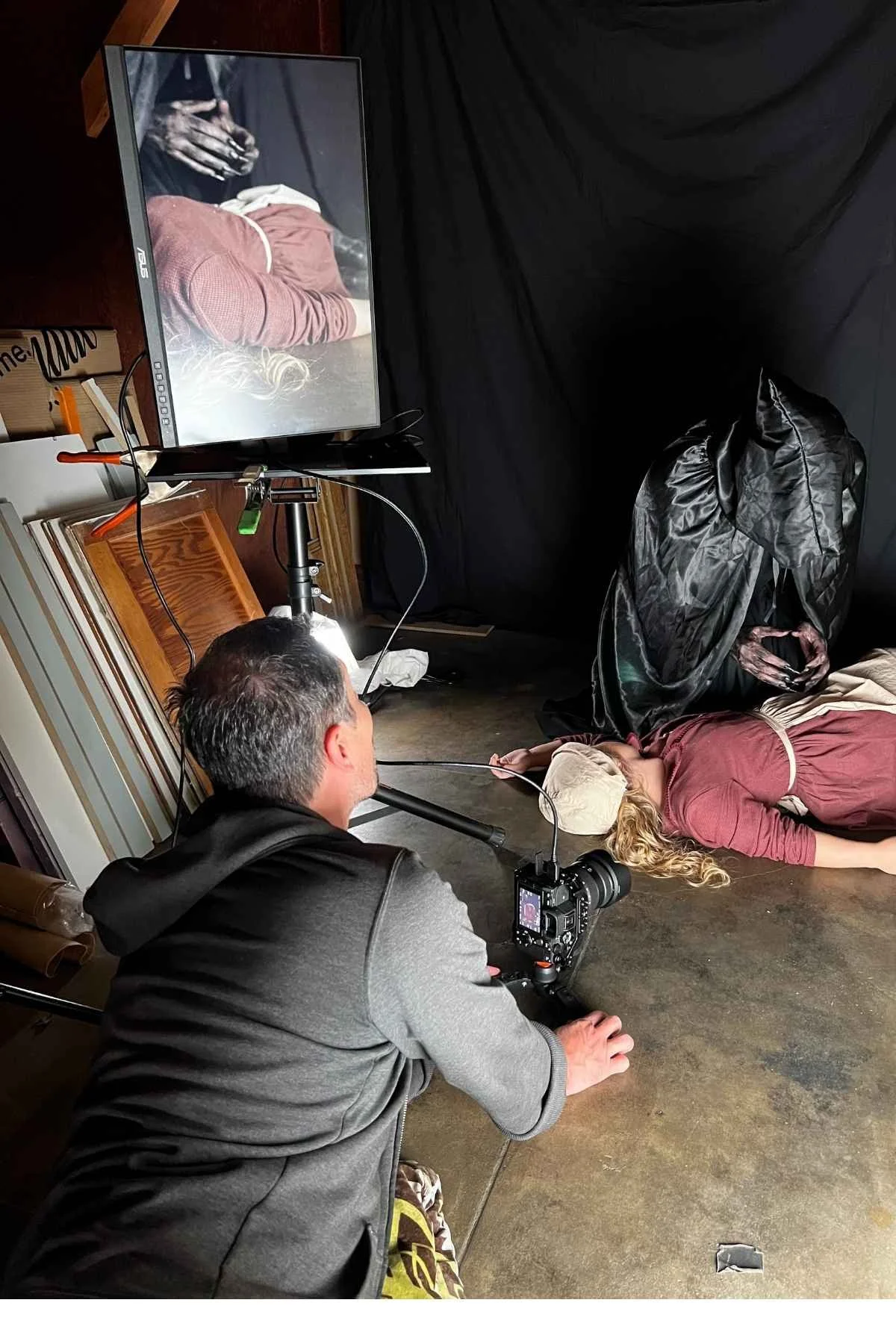 A behind-the-scenes shot of a photo shoot where a woman with long blonde hair is lying on the floor and a photographer is taking her picture. The woman is wearing a beige cap and a reddish shirt. A monitor nearby displays the image being captured, and there are various photographic equipment, including a camera, tripod, and lighting, in the scene.