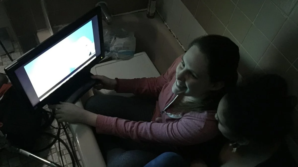 Two women sitting together in a dimly lit room, looking at a computer monitor that displays a cartoon landscape with clouds. One woman is smiling, and they are both engaged with the screen.