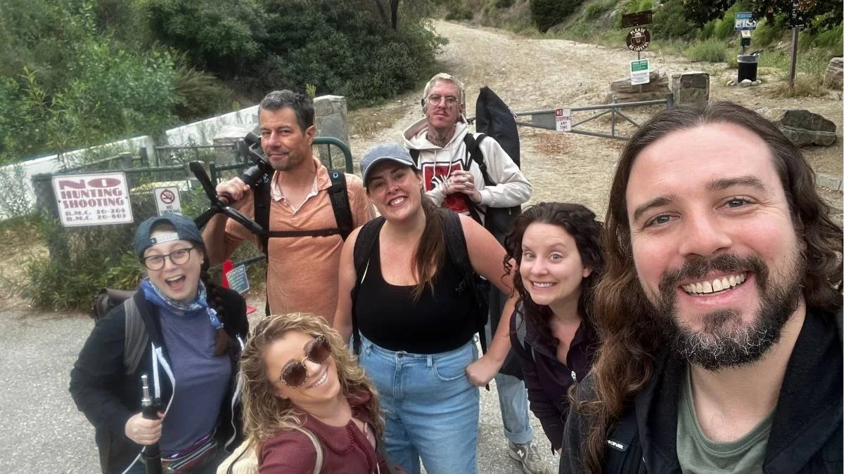 Group of six smiling people taking a selfie outdoors on a hiking trail with trees and signs in the background.