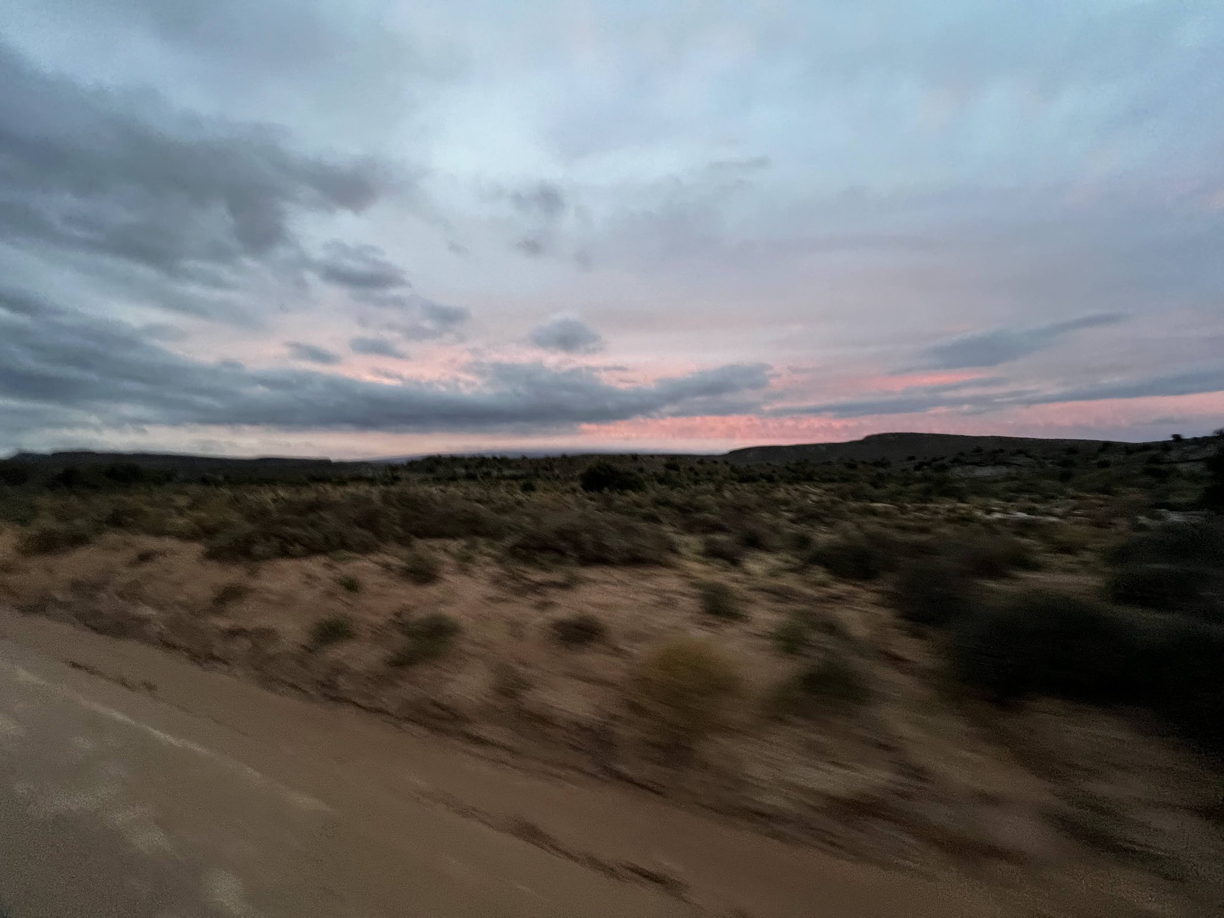 A blurred photo of a desert landscape at sunset, with a gravel road in the foreground and low rolling hills under a partly cloudy sky with pink and blue hues.