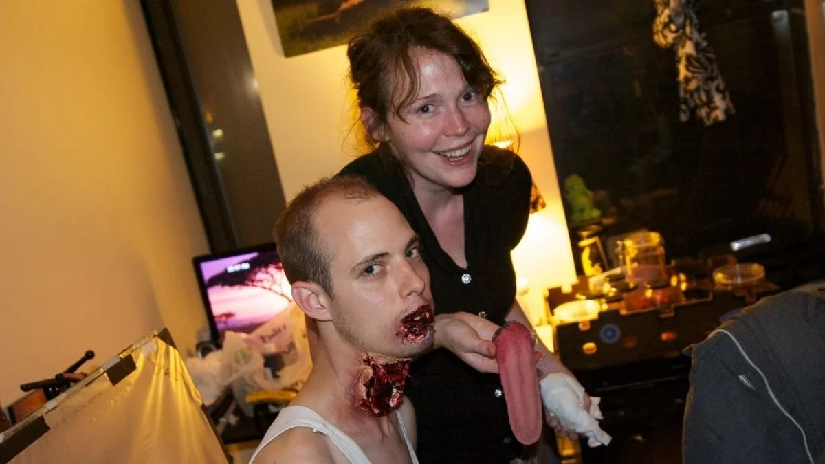 A woman with curly hair smiling and a man with fake wounds on his neck and mouth, holding a realistic-looking fake severed tongue, in a cluttered room with a television and various household items.