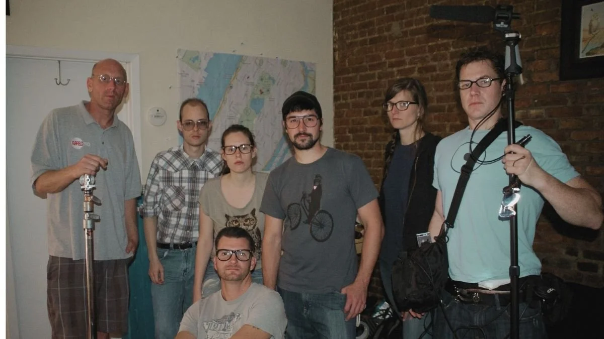 Group of six people posing indoors, with one person seated on the floor, and a brick wall in the background. Some individuals are holding camera or lighting equipment.