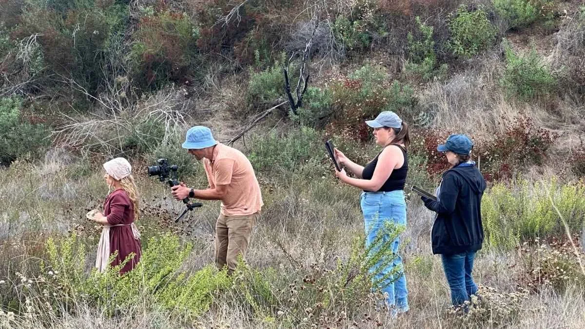 Four people outdoors in a gravel and brush landscape, one girl in a maroon dress and hat, a man with a camera, and two women, one with a tablet and one with a notepad.
