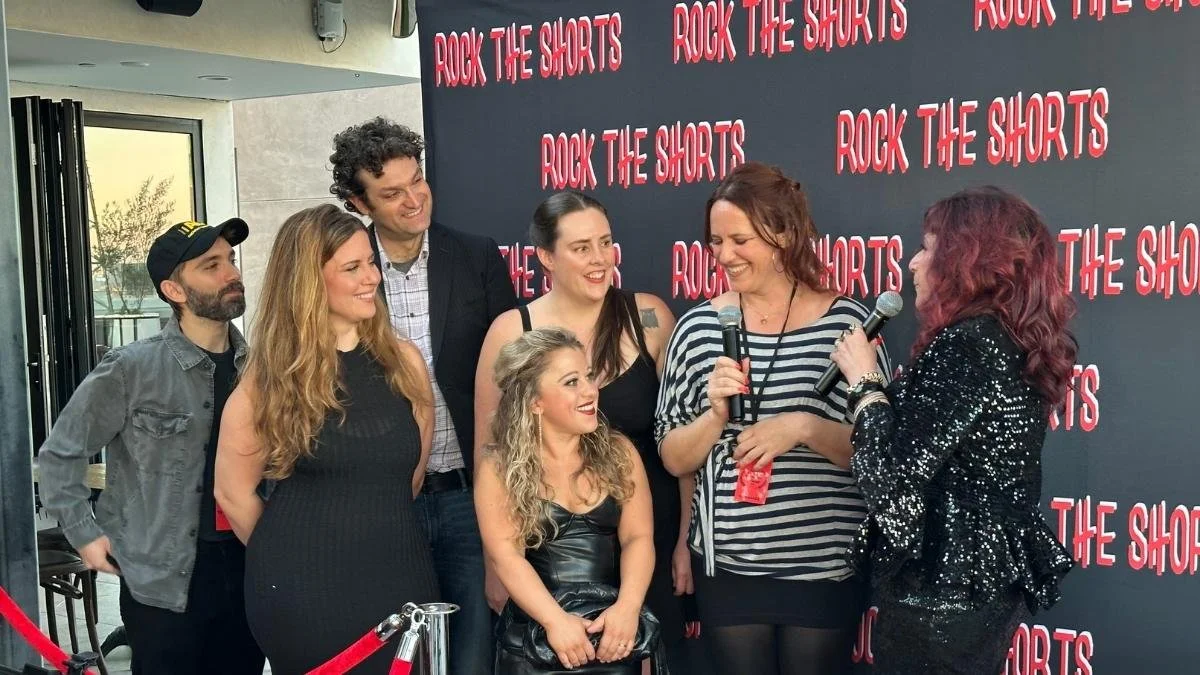 Group of six people, five women and one man, standing in front of a black backdrop with red text that reads 'Rock the Shorts'. Two women are holding microphones and talking, and everyone appears to be smiling and engaged in a conversation.
