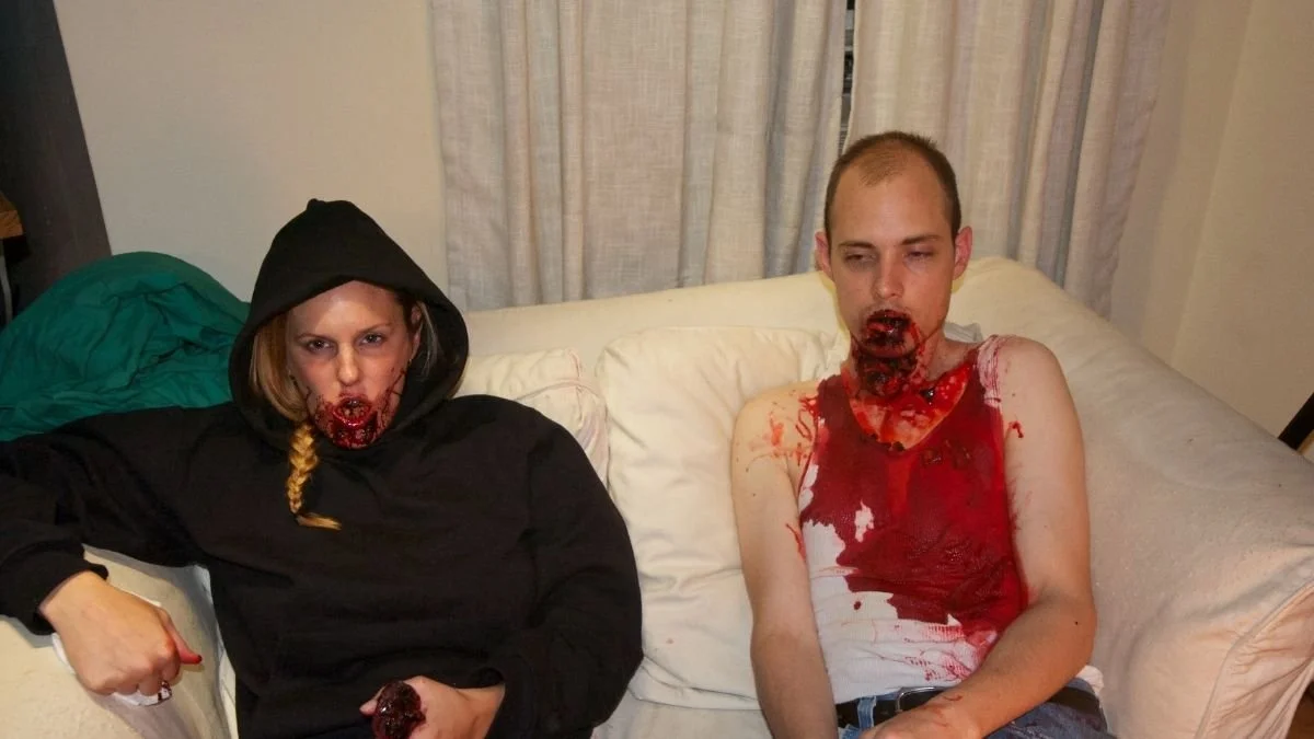Two people sitting on a couch with wounds and fake blood on their faces and clothes, dressed in Halloween costumes, in a room with curtains in the background.