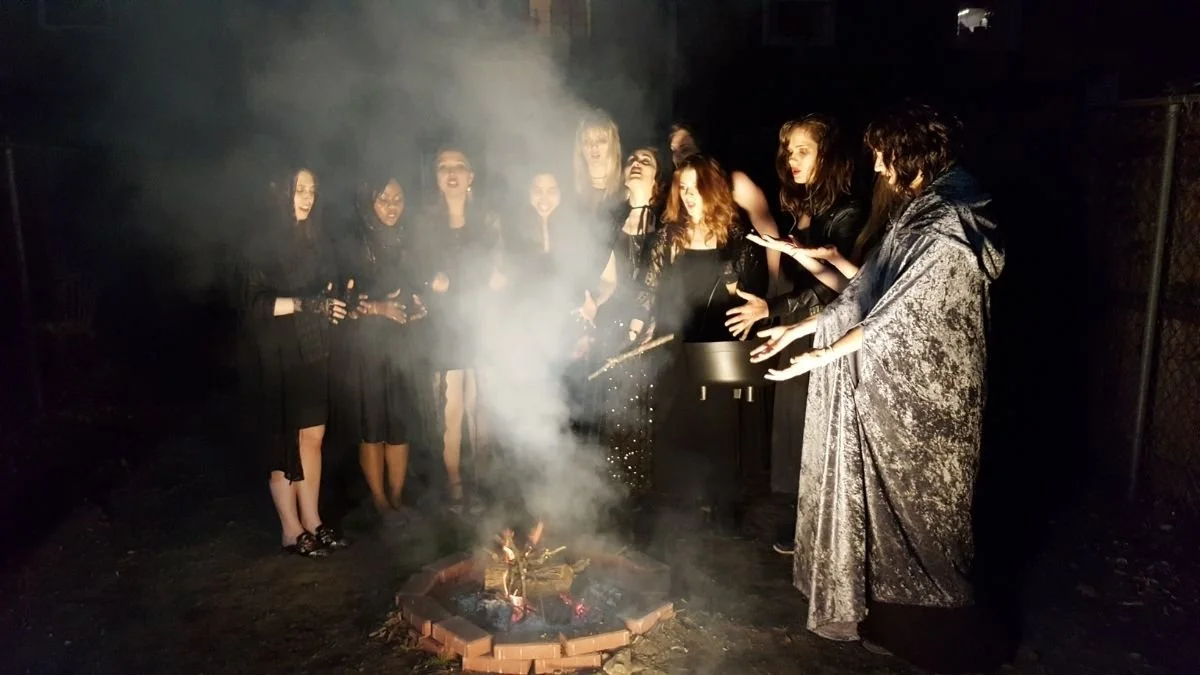 Group of women in black costumes gathered around a small outdoor fire on a dark night, celebrating with sparklers and drinks.