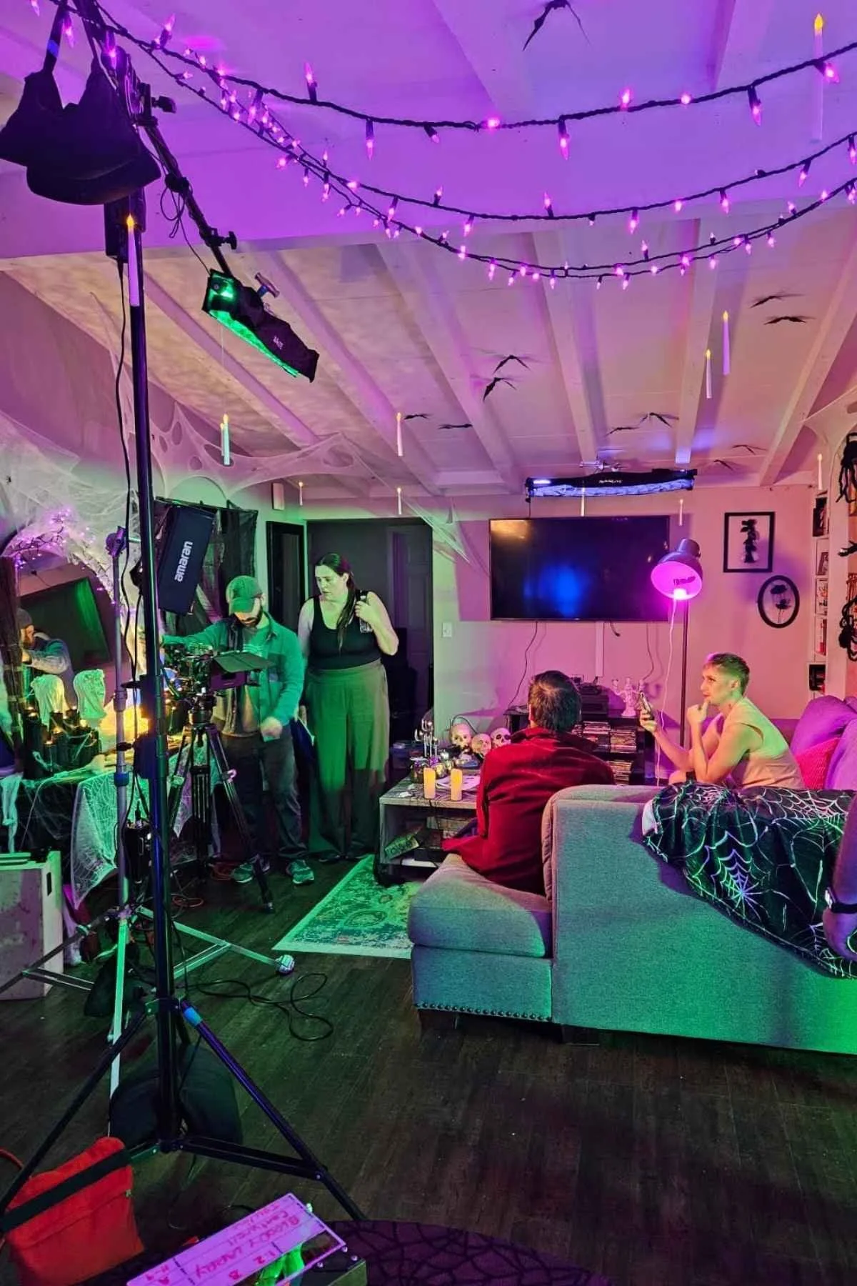 Living room decorated with purple string lights and Halloween decorations, featuring people preparing for filming or streaming, with a large TV, a sofa, and spooky props on the table.