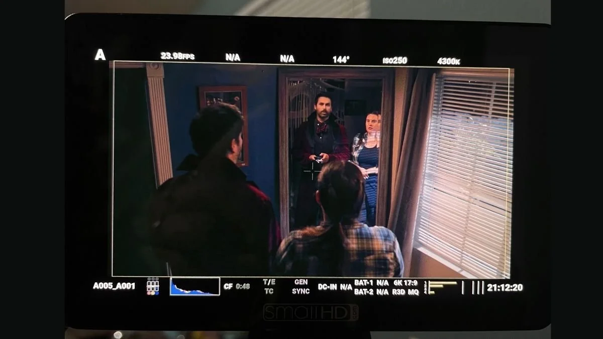 People standing in a dimly lit room, visible through a camera monitor, with blinds on a window on the right side.