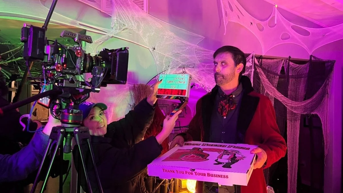 A man with a beard in a red jacket holding a pizza box, standing in front of a camera on a tripod, with a woman next to him holding a clapperboard. The scene is decorated with spider web themed decorations and colorful lighting.