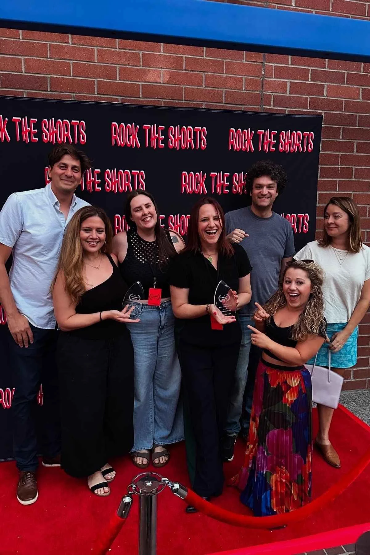 A group of seven people standing on a red carpet in front of a black backdrop with red text that says, 'ROCK THE SHORTS.' They are smiling and holding glass awards. The group includes six women and one man, dressed casually, at an event or celebration.