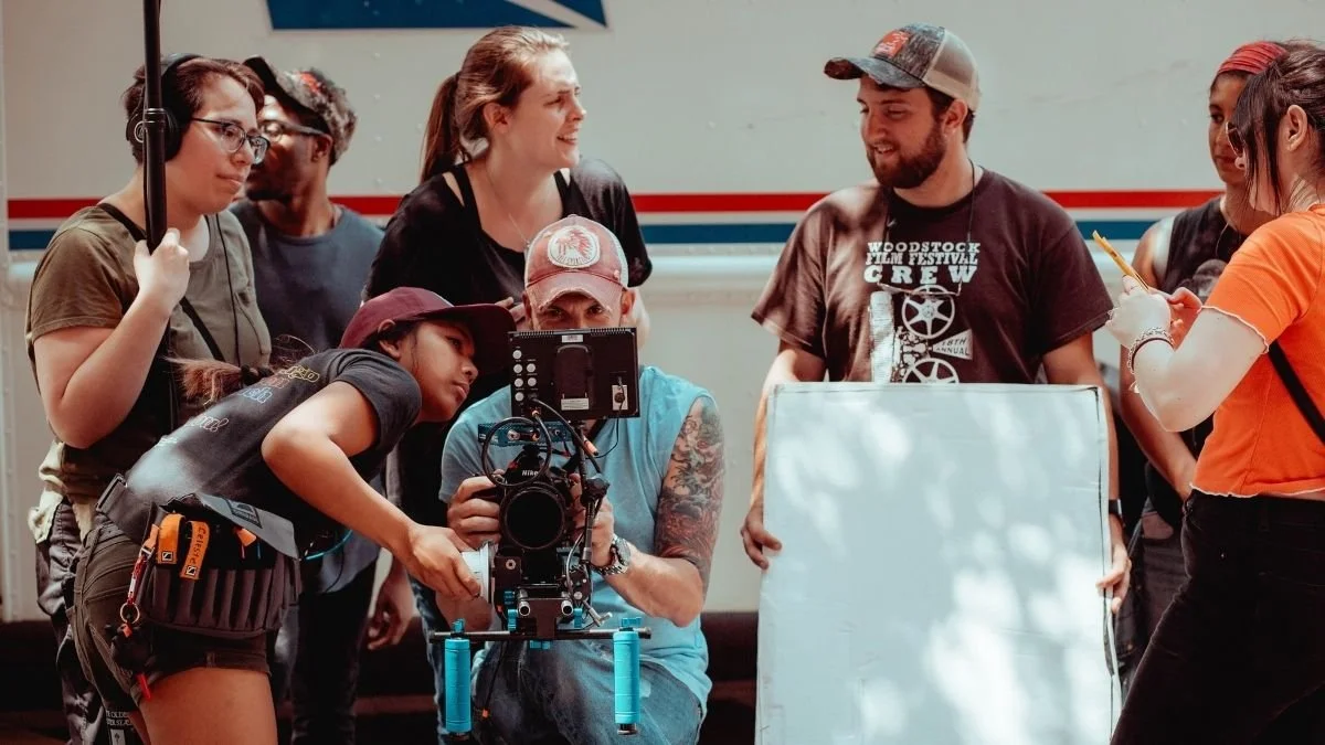 A group of people working on a film set, with a camera operator adjusting a camera while others hold equipment and discuss.