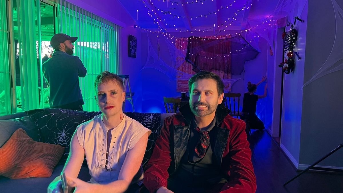 People dressed in Halloween costumes sitting on a couch, with Halloween decorations and purple and green lighting in the background.