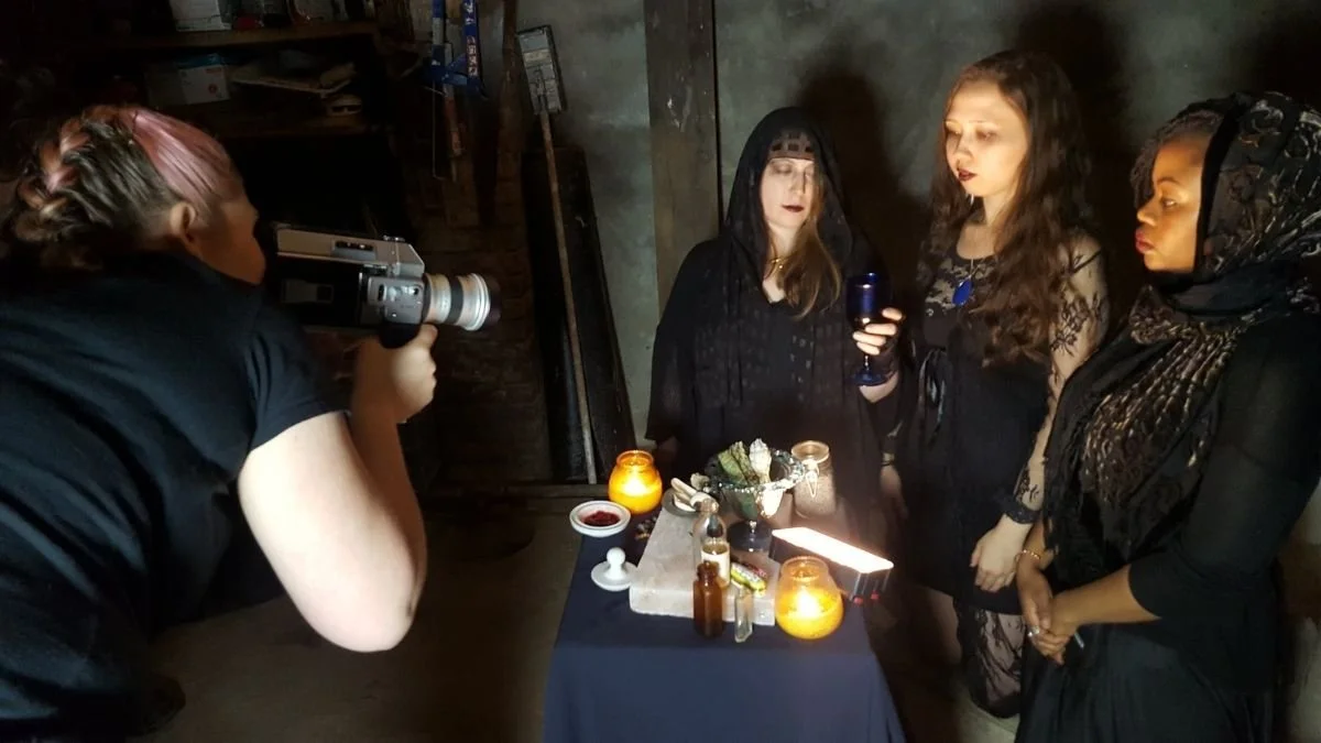 Three women in black and dark clothing standing at a table with candles and potion-like bottles, being photographed by a woman with pink hair using a camera in a dimly lit room.