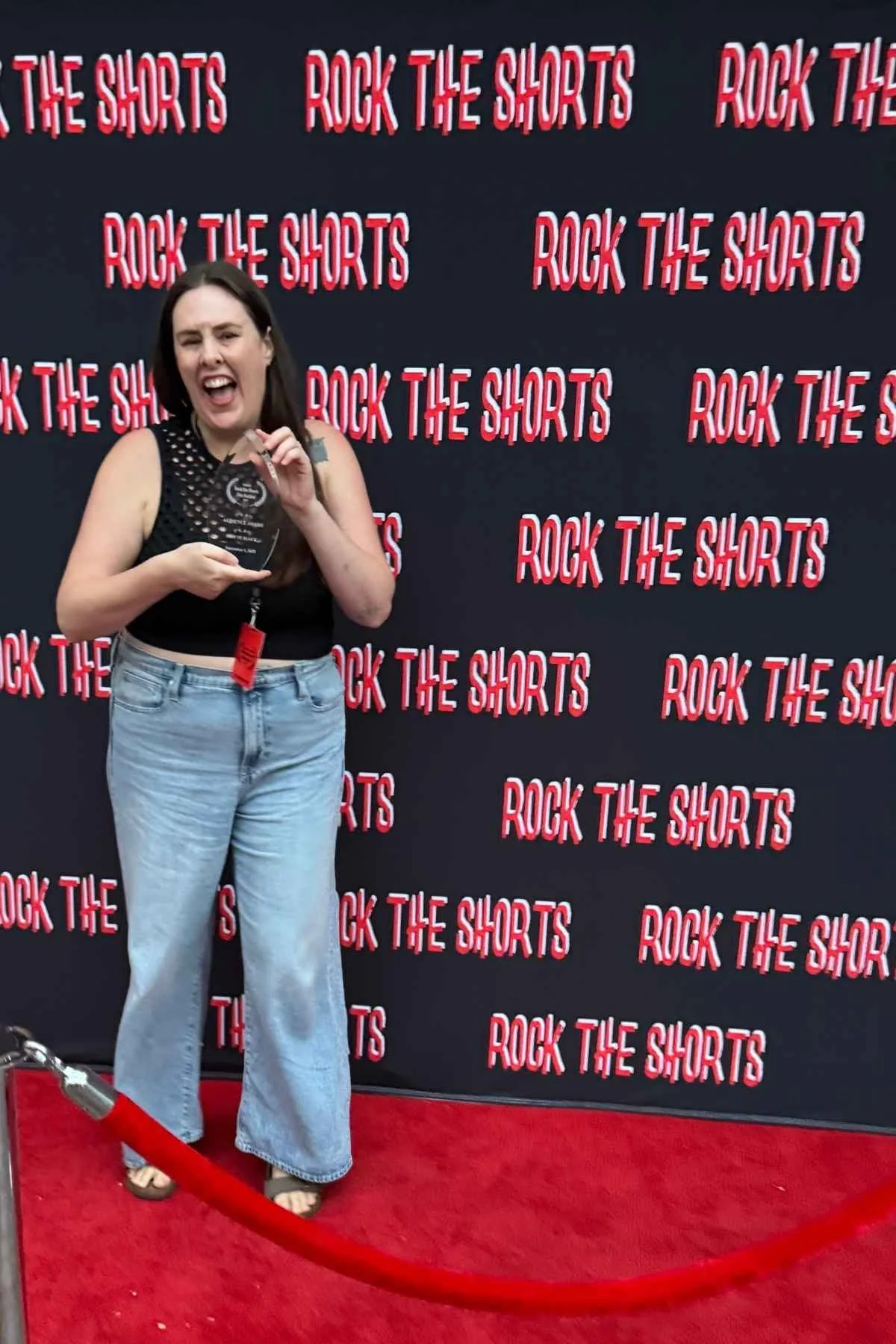 Woman celebrating on a red carpet holding an award, standing in front of a backdrop that says 'Rock the Shorts' repeatedly.