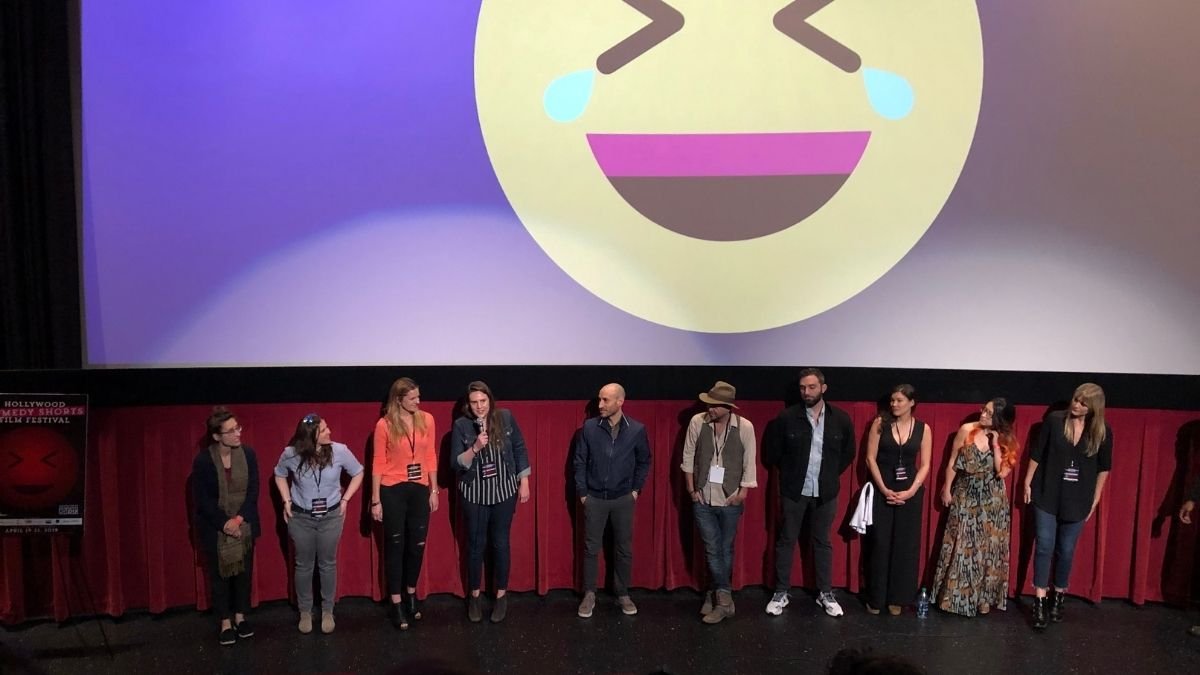 Group of people standing on stage in front of a large screen displaying a laughing emoji with tears.