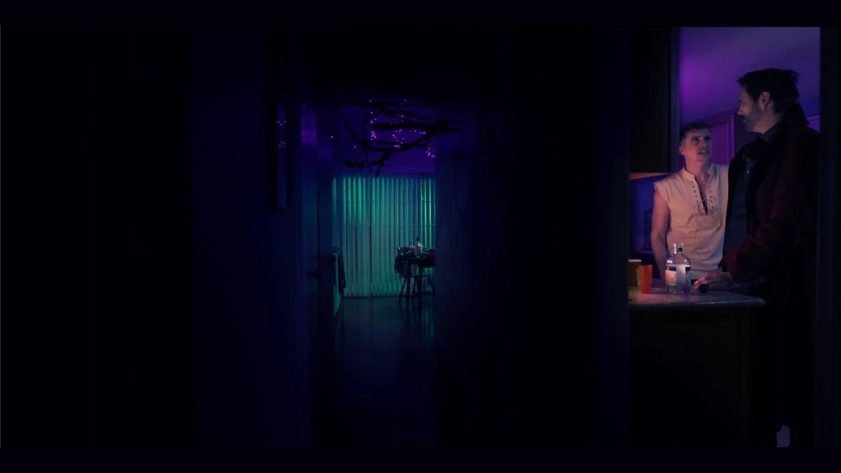 Two people standing and talking in a dimly lit room with neon lighting, kitchen counter with bottles and cups.