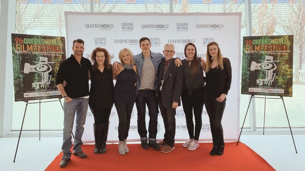 A group of seven people standing on a red carpet at the Queens World Film Festival, posing for a photo in front of a white backdrop with the festival's logo. Two posters for the festival are placed on easels on either side of the group.