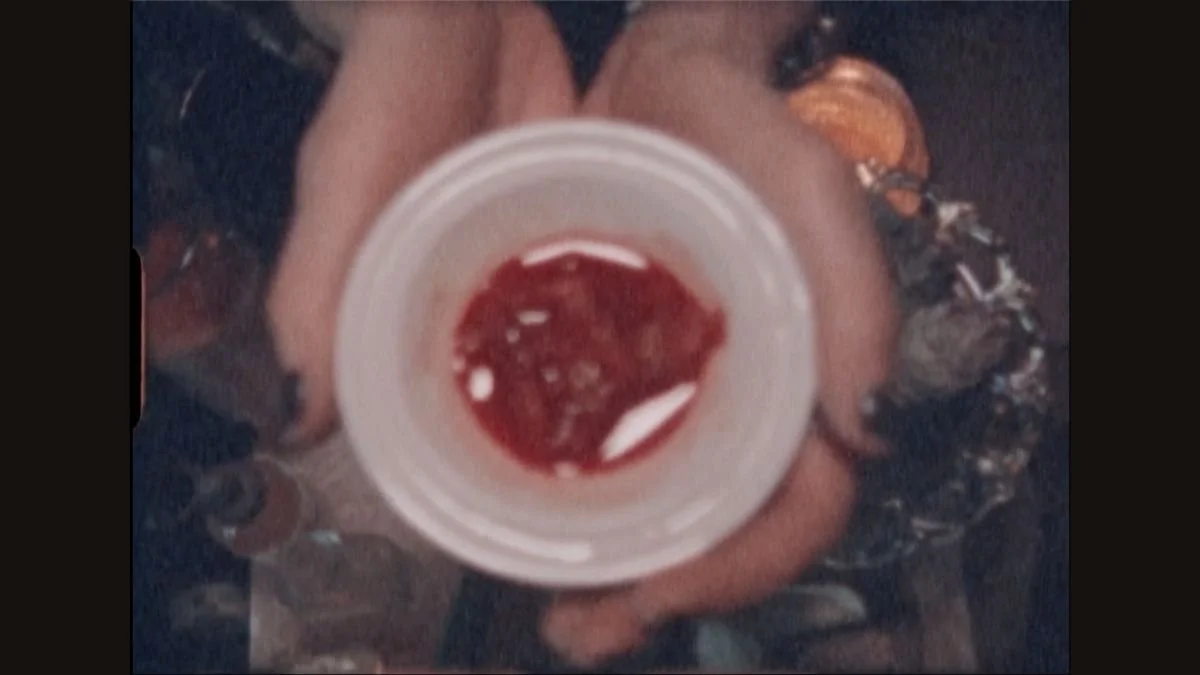 Person holding a cup filled with a red beverage, with fingers visible around the cup, and wearing a dark, patterned outfit.