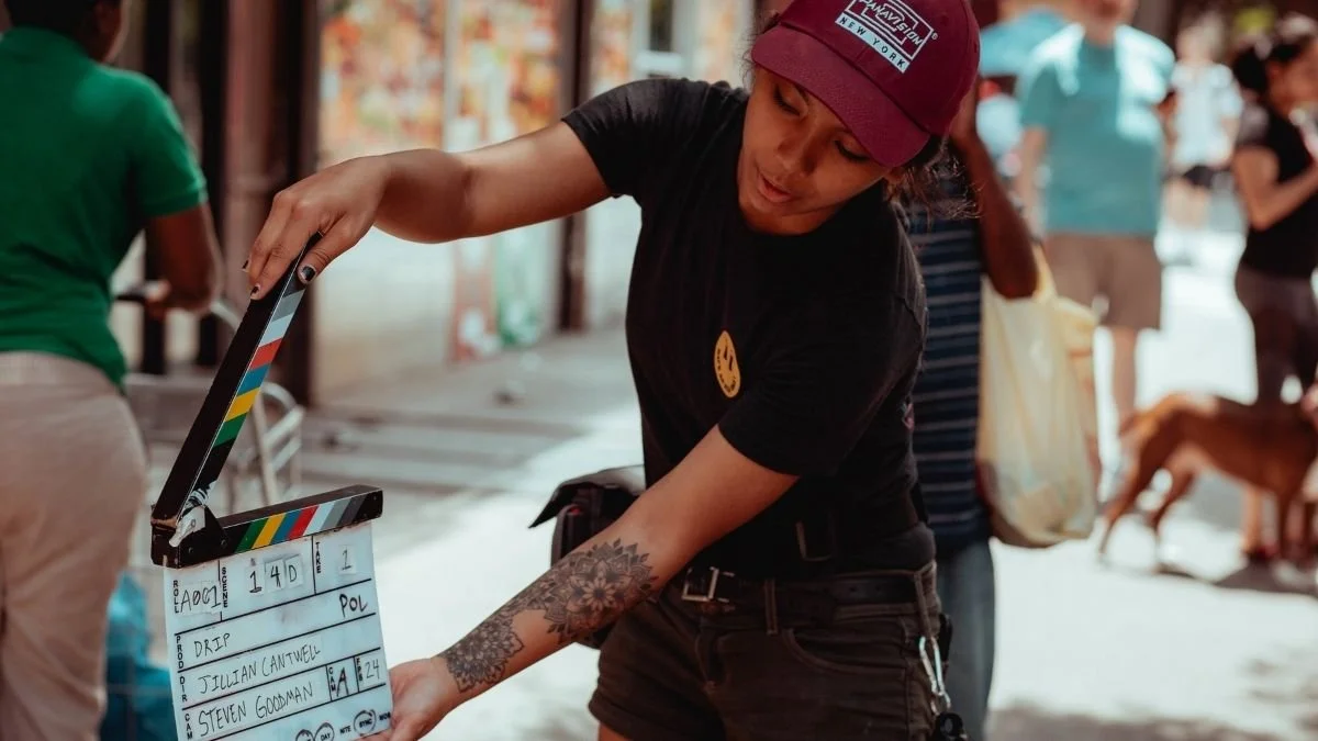 A woman with tattoos on her arm and wearing a maroon cap and black T-shirt is working on a film clapboard on a busy street, with people and a dog in the background.