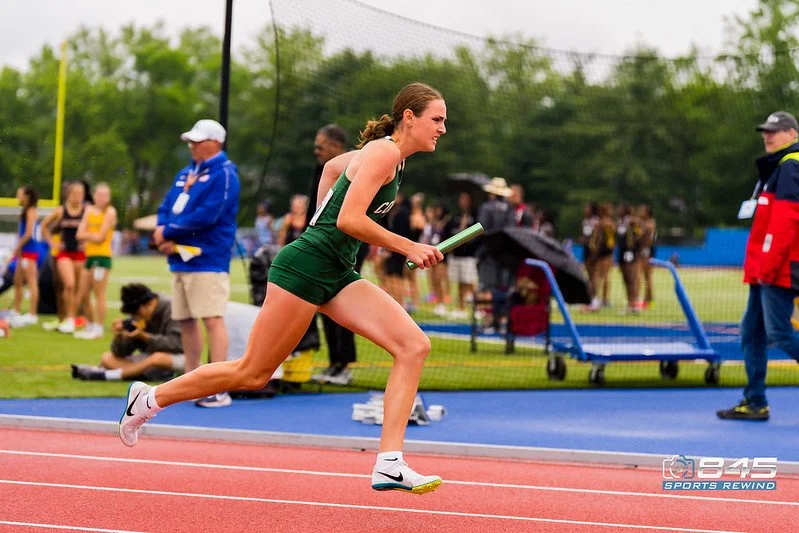 A Cornwall High school female athlete in green running shorts and a tank top competing in a relay race, holding a baton on an outdoor track, with spectators and officials in the background.
