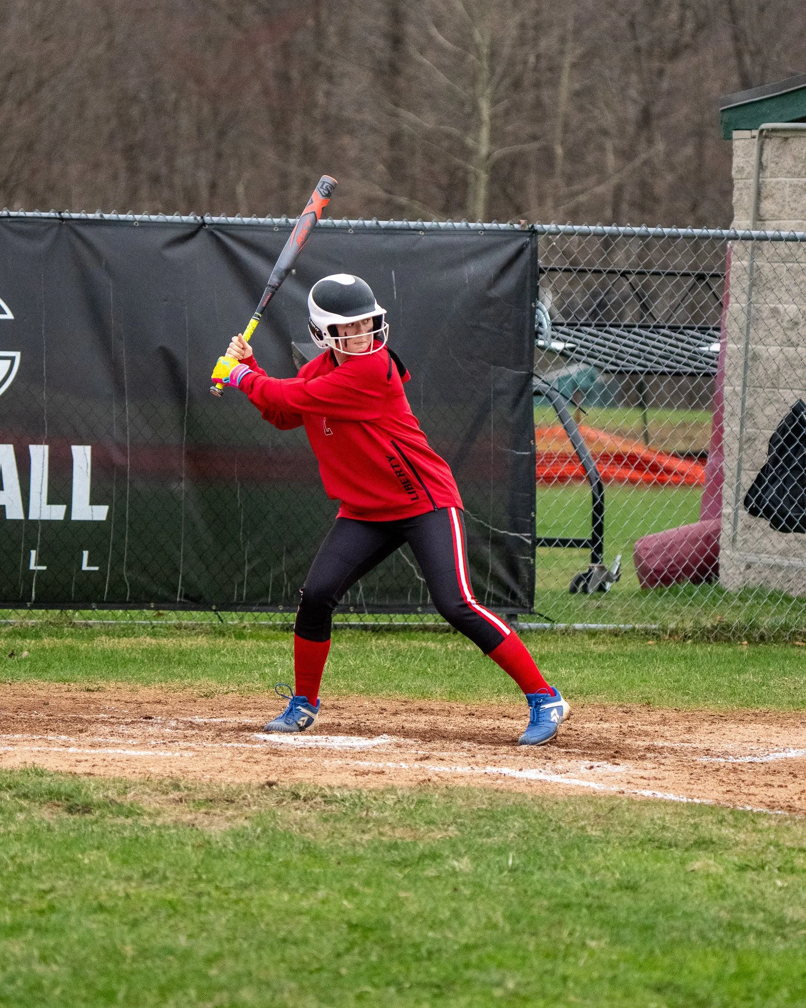 A Liberty High Schoolfemale softball player stands at the batting box, about to swing a bat during a game on a dirt field.
